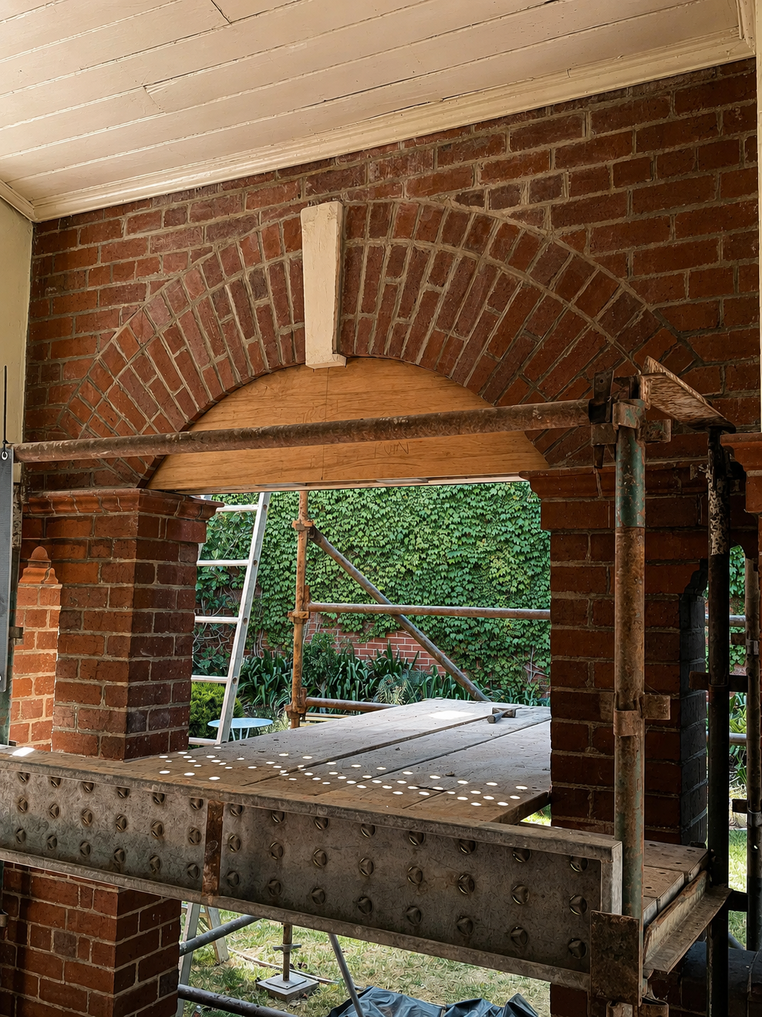 Construction site showing a renovated heritage brick archway with scaffold and ladder outside, green foliage in the background.