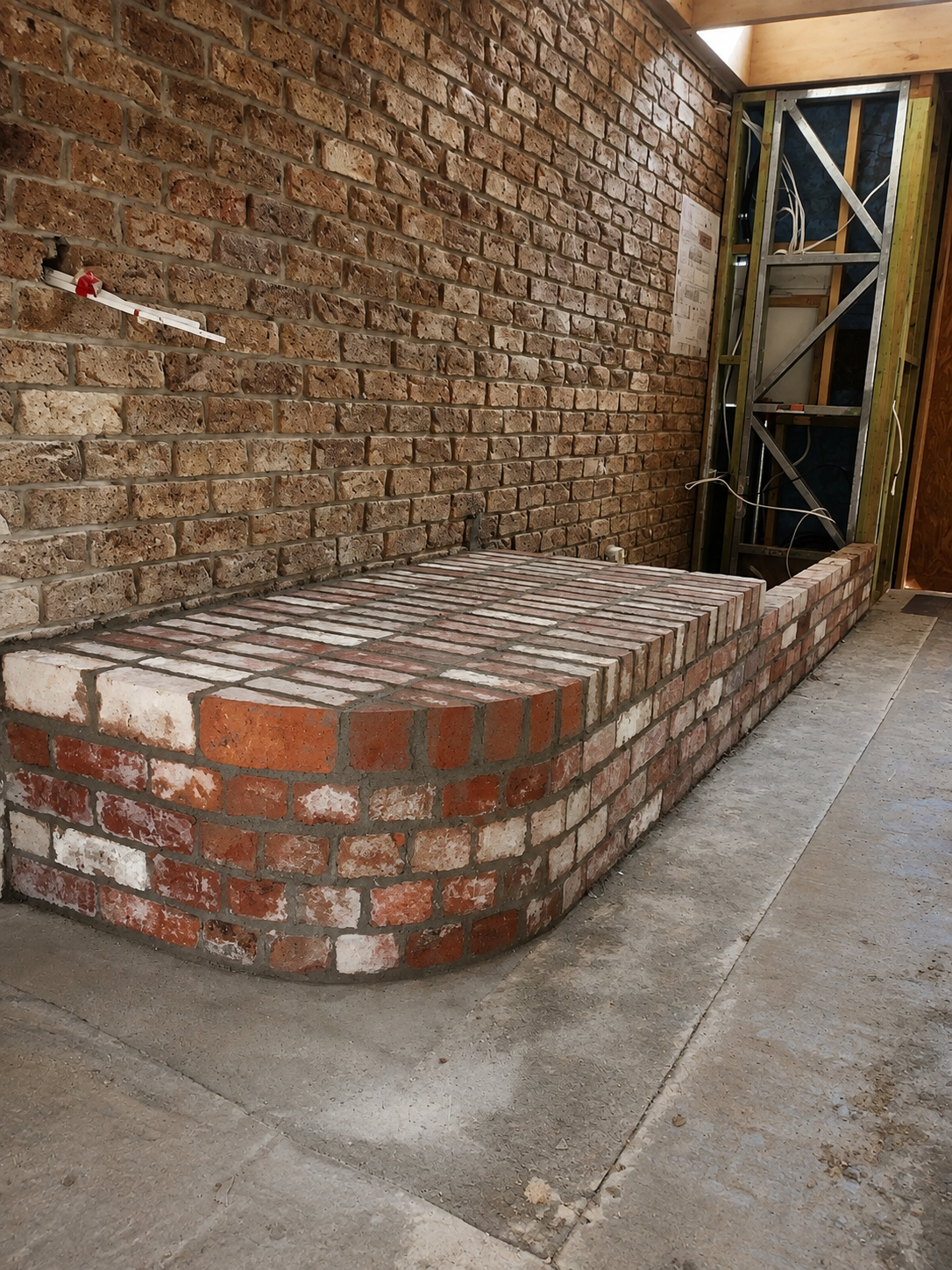 A recycled red brick bench under construction with a curved wall and an unfinished space for a door or window, showing construction framing and electrical wiring.