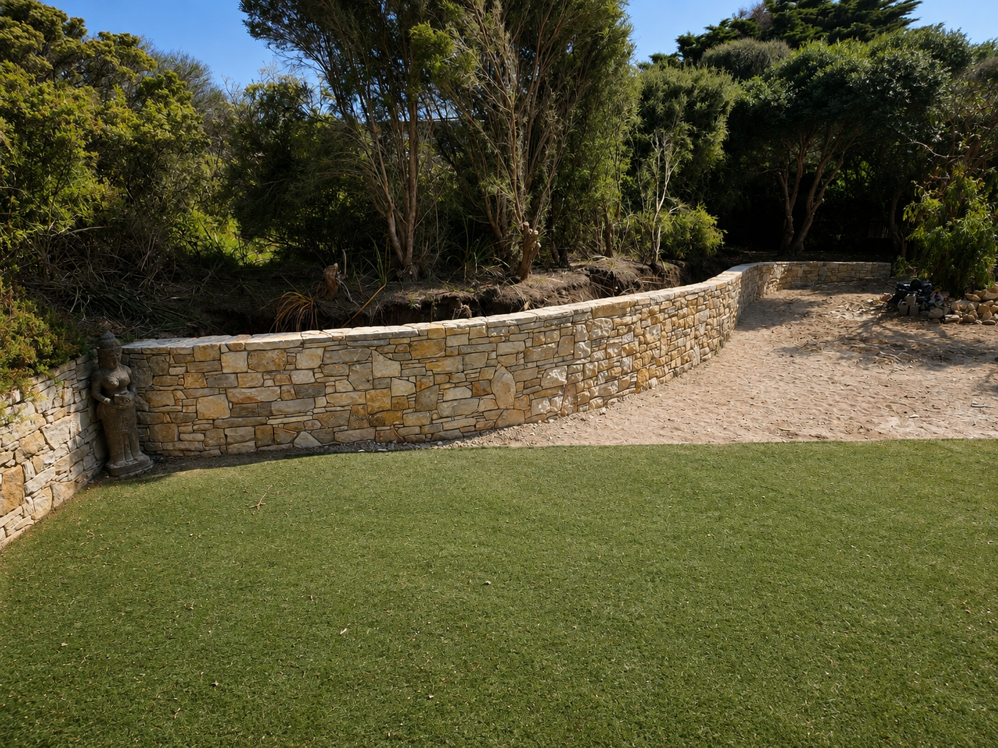A stone retaining wall, separating a grassy lawn from a dirt and sand pathway with trees and bushes in the background.