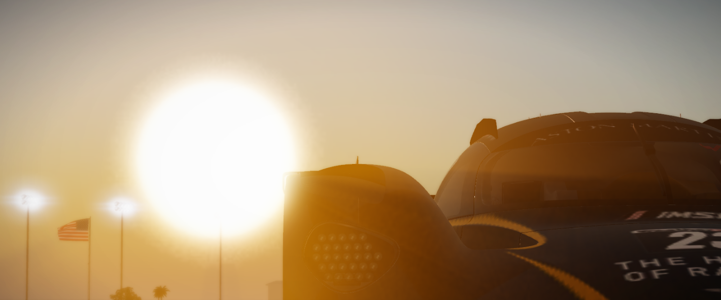 The image shows a race car with the sun setting or rising in the background. There are multiple American flags on flagpoles and stadium lights behind the car.