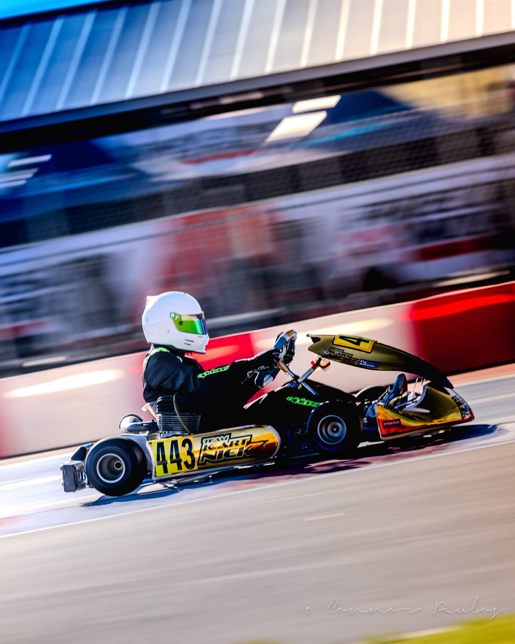 A go-kart racer wearing a white helmet and black racing suit driving on a track with a blurred background.