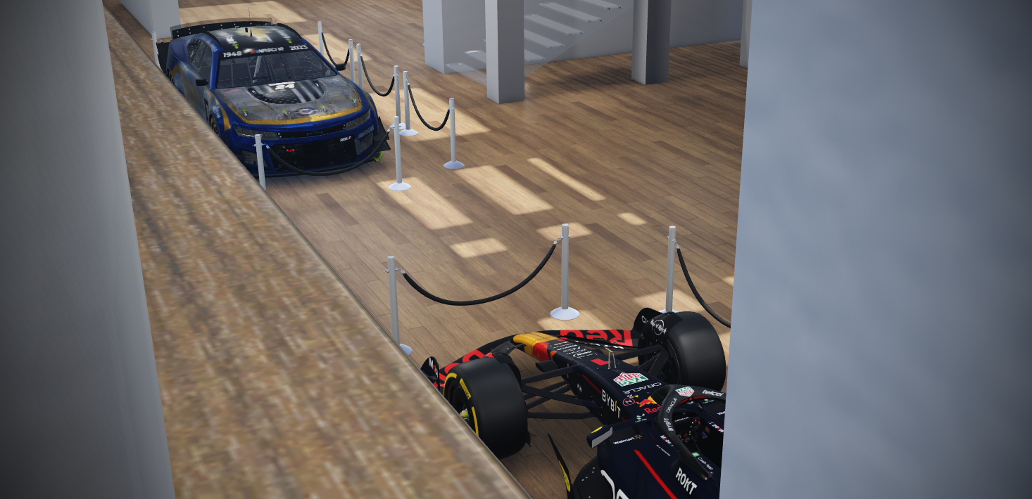Two race cars on display inside a museum behind protective stanchions with black ropes, viewed through a gap between a brick wall and a white wall.