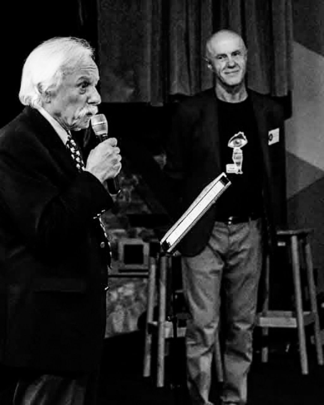 Two elderly men are standing on a stage, one reading from a book and holding a microphone, the other listening with a slight smile, set against a dark curtain.