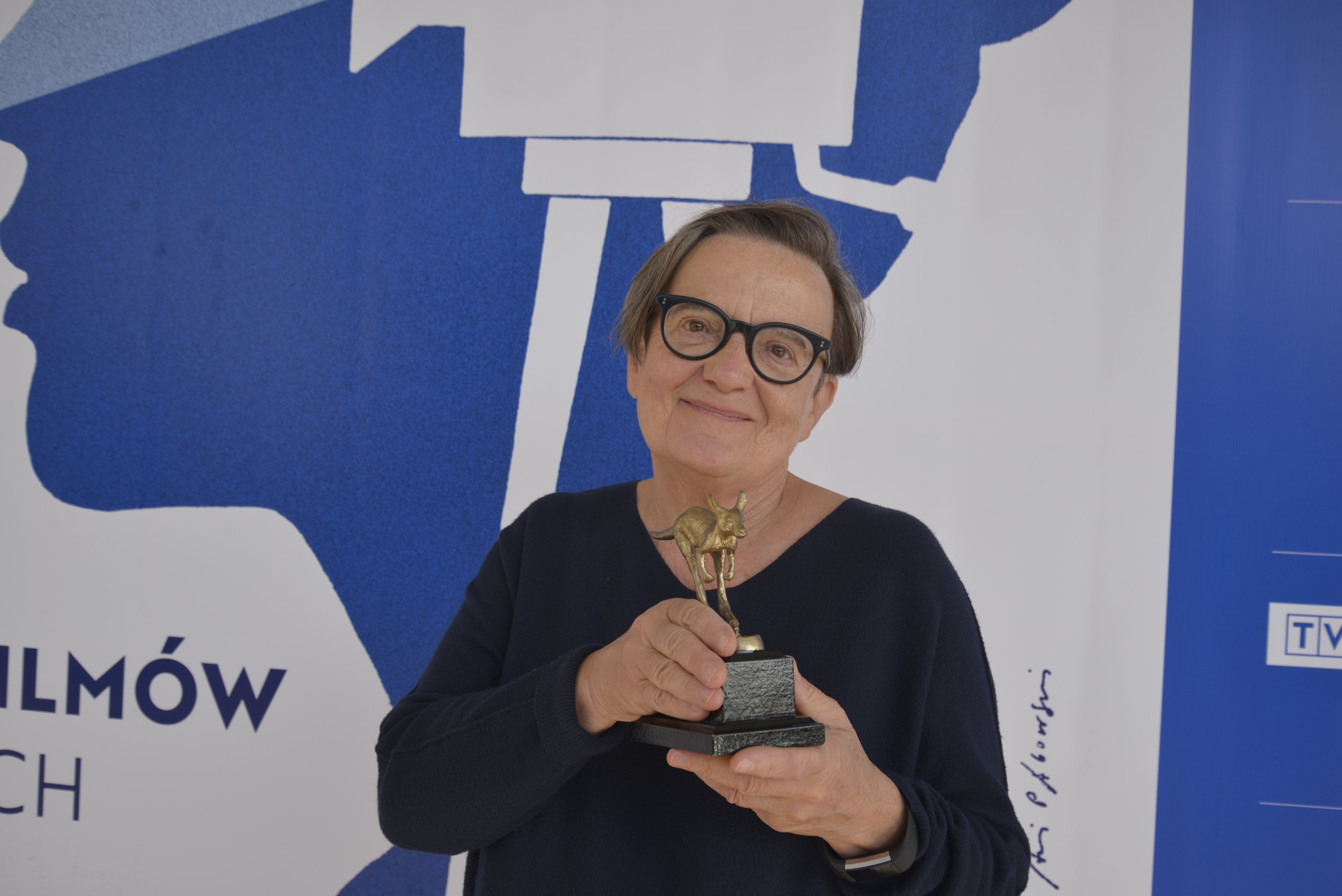 A woman with short hair and glasses smiling and holding a trophy shaped like a bull in front of a blue and white mural.