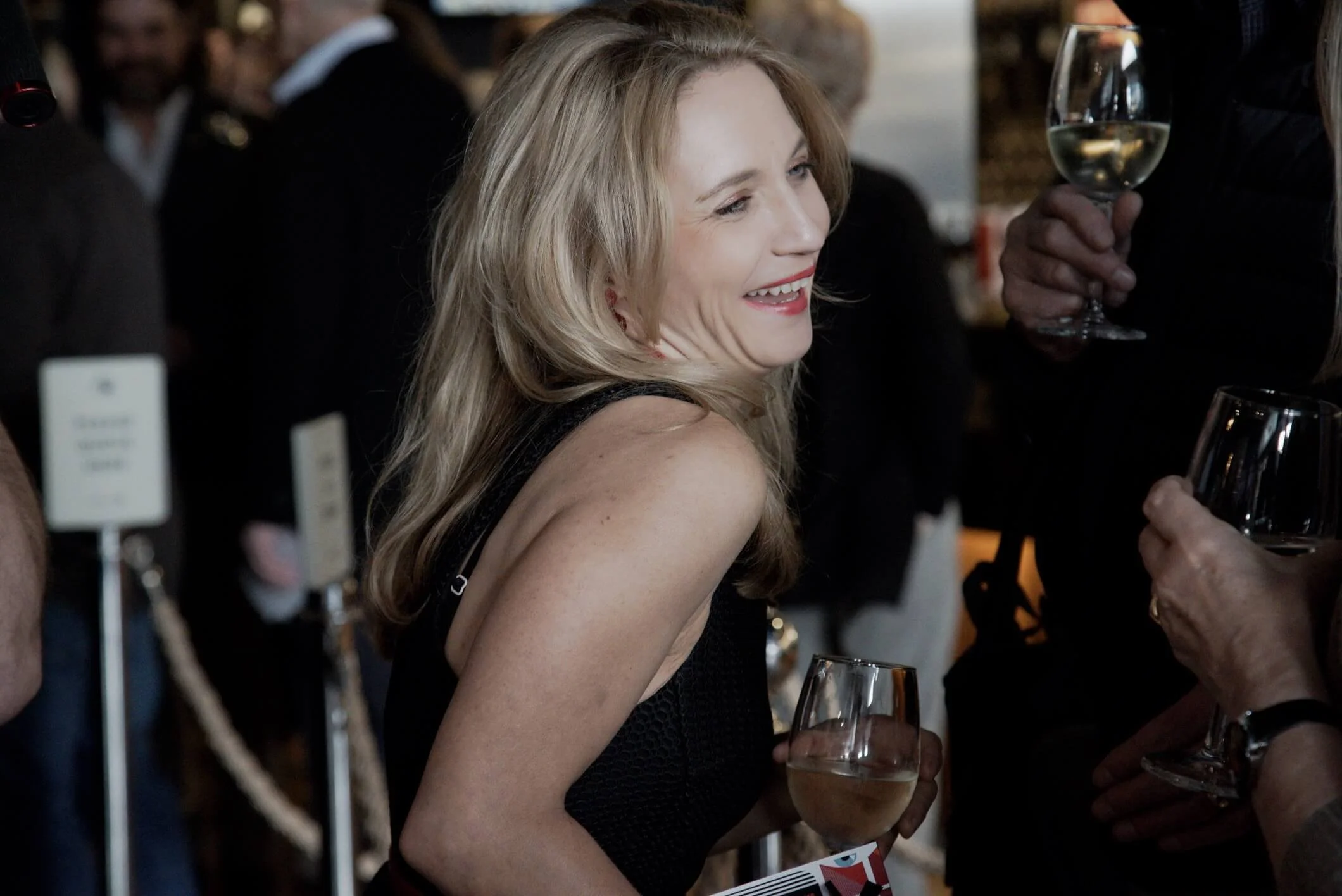 A woman with long blonde hair smiling and holding a glass of white wine at a social gathering, surrounded by other people.