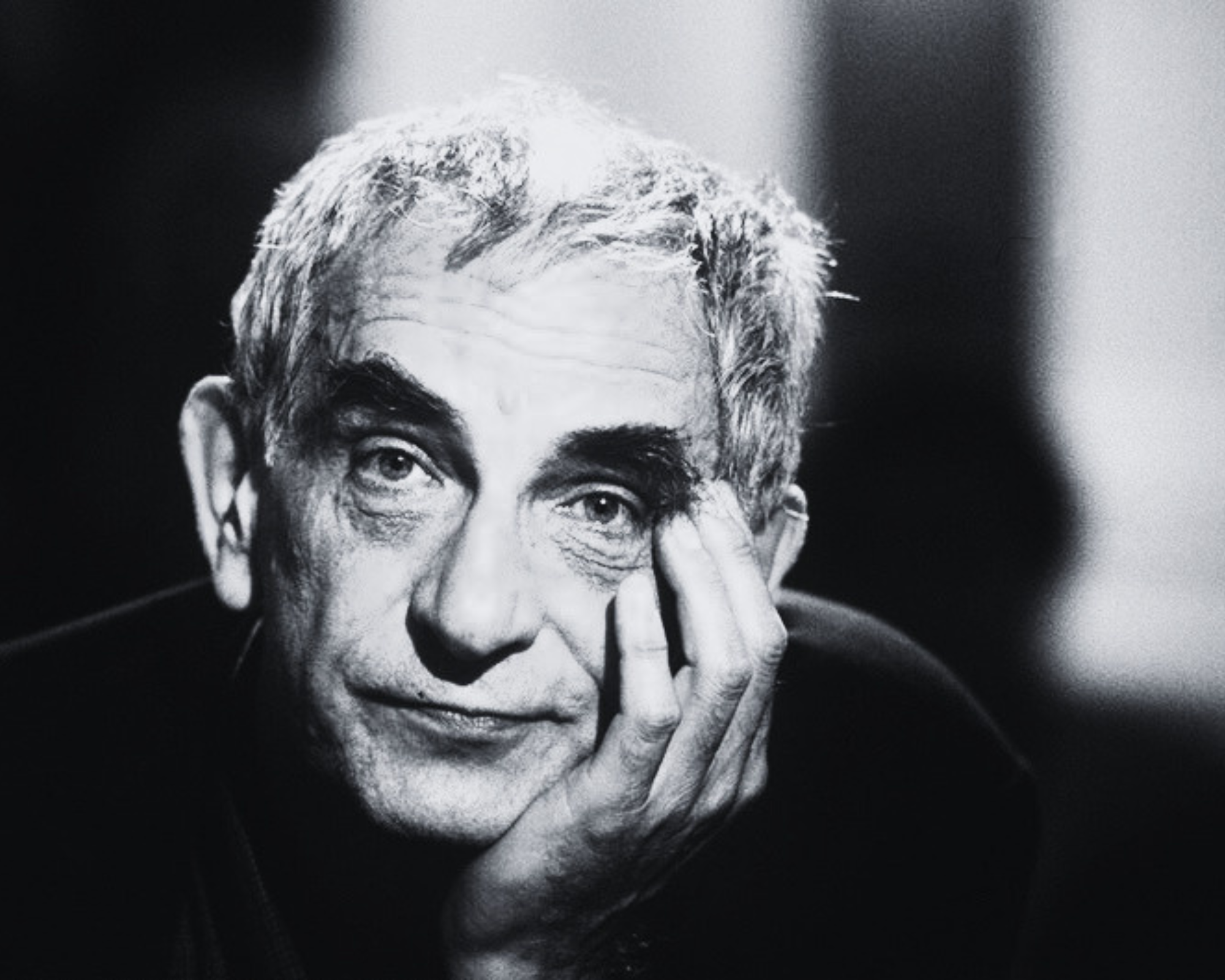 Close-up black-and-white portrait of an elderly man with glasses, smiling, making a peace sign with his fingers.