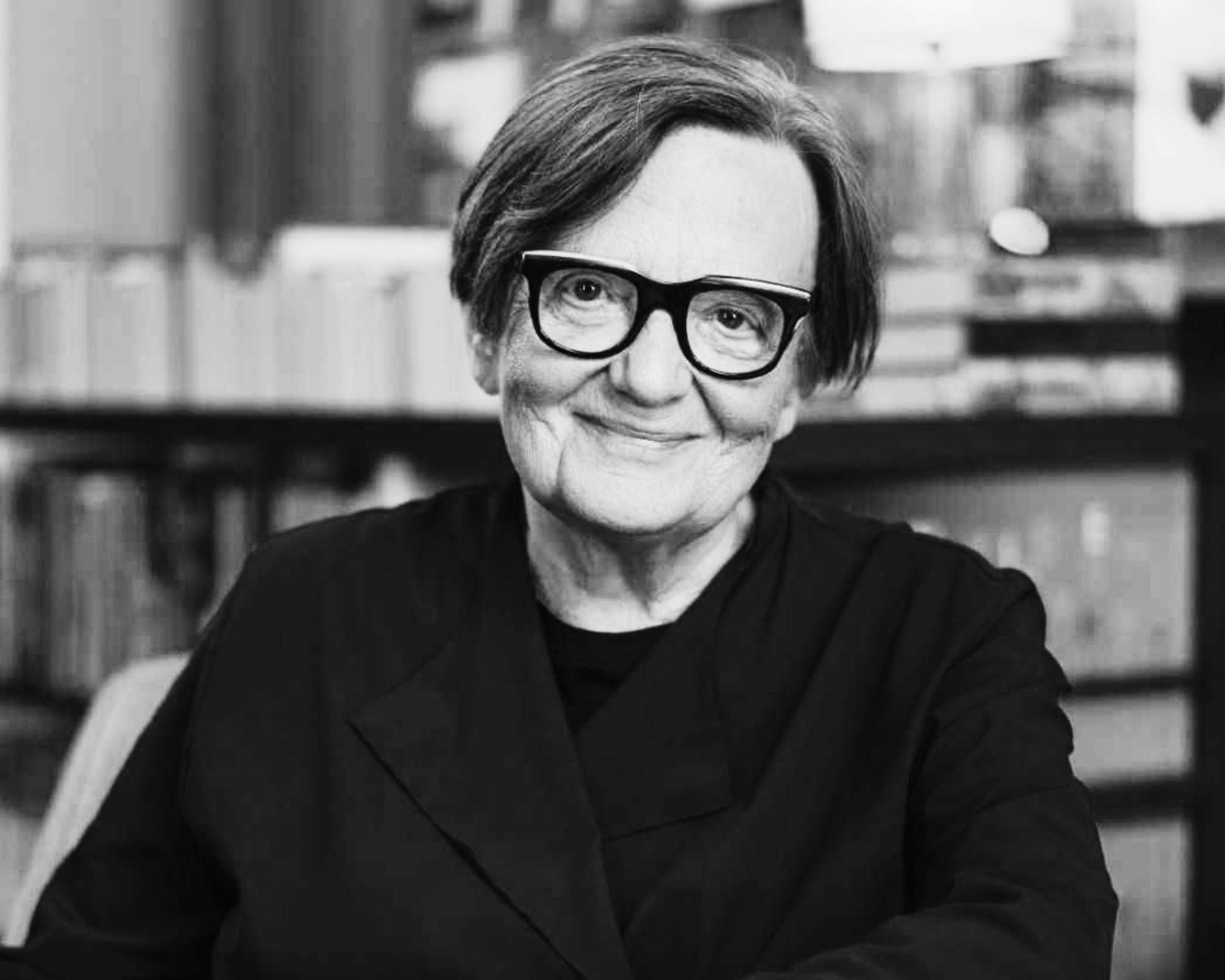 A smiling woman wearing glasses, with short hair, in a bookstore or library setting.