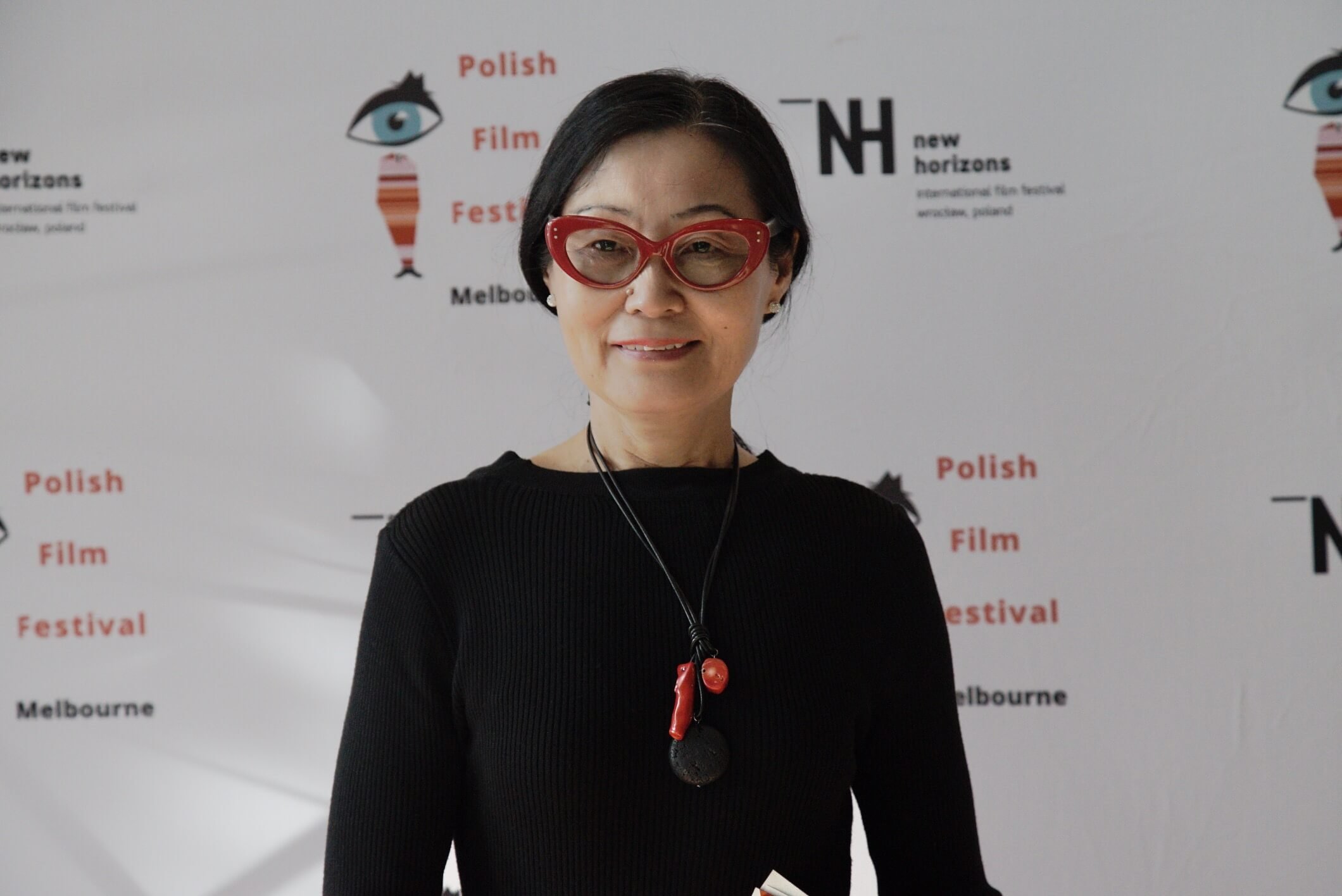 A woman wearing red glasses with a black frame, a black top, and a red and black necklace, standing in front of a white background with logos and text related to a film festival in Melbourne.