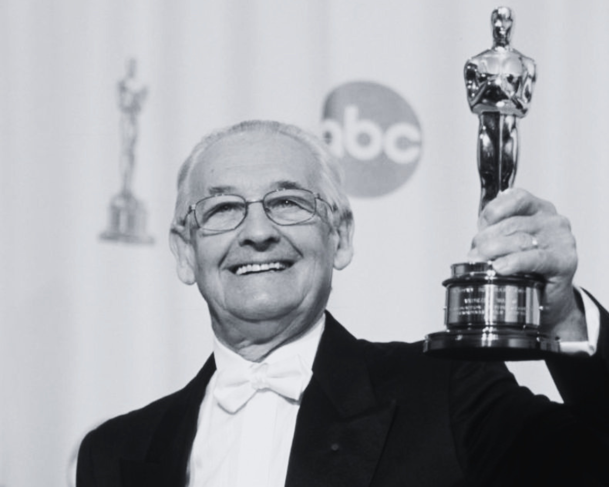 A smiling elderly man wearing glasses in a tuxedo holding an Oscar trophy.
