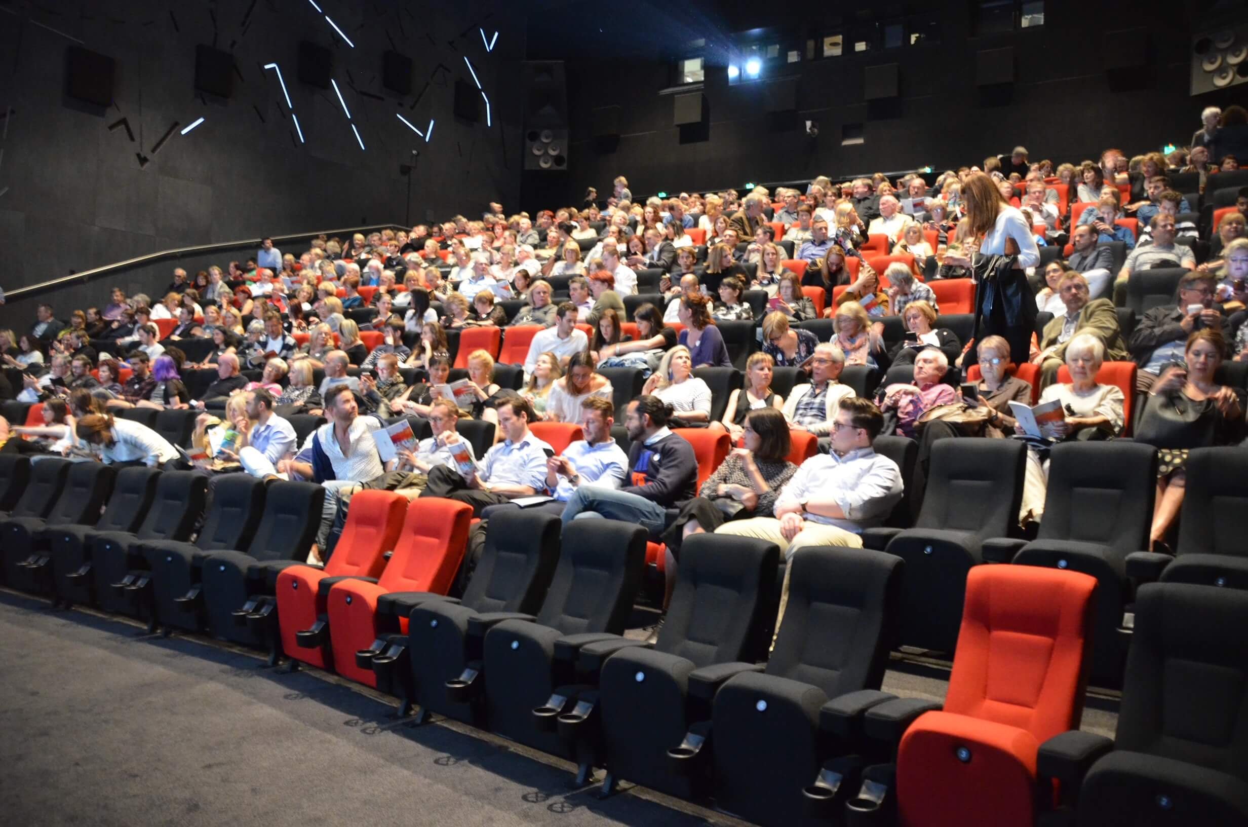 A large auditorium filled with people seated in red and black chairs, attending an event or presentation. Some are reading, using phones, or talking, with a dark wall and geometric light design in the background.