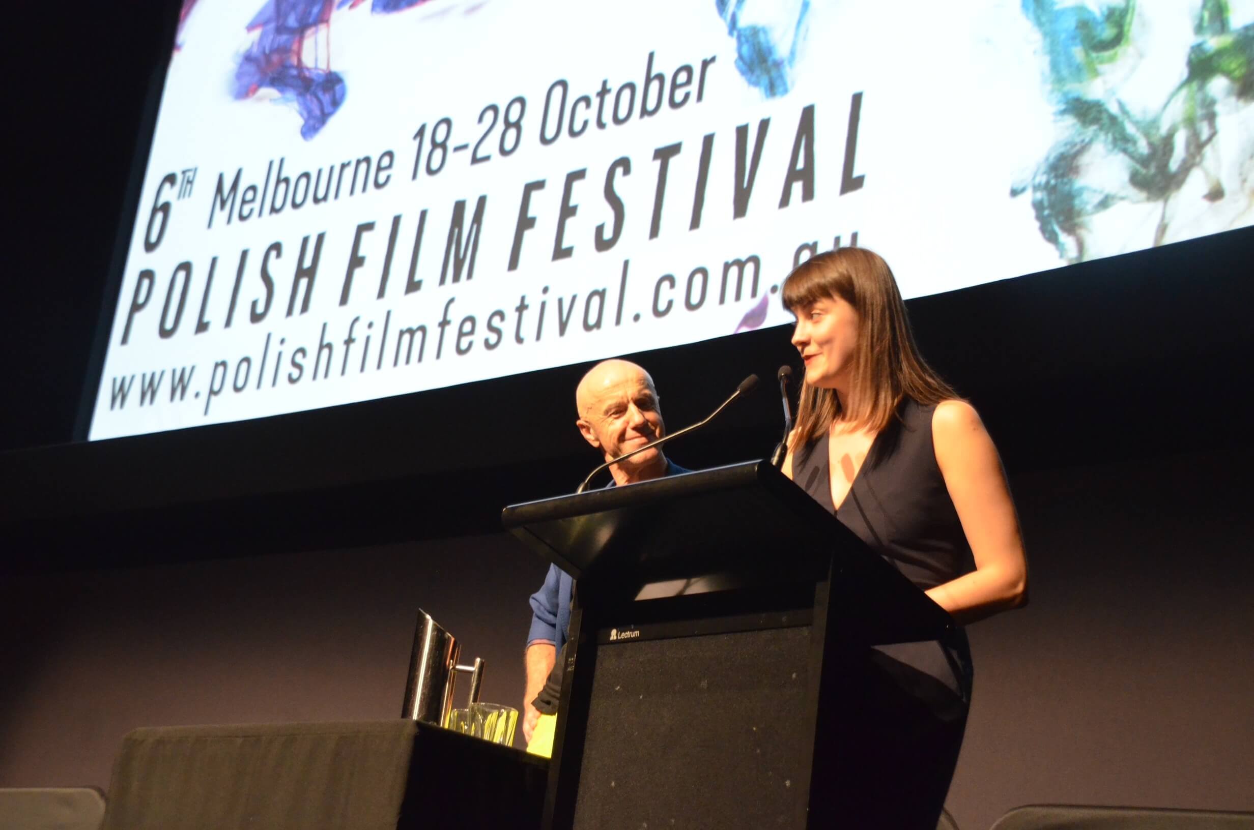 Two people on stage at the 6th Melbourne Polish Film Festival, with a large banner displaying event details behind them. One woman is speaking at a black lectern, and a man is standing beside her, smiling.