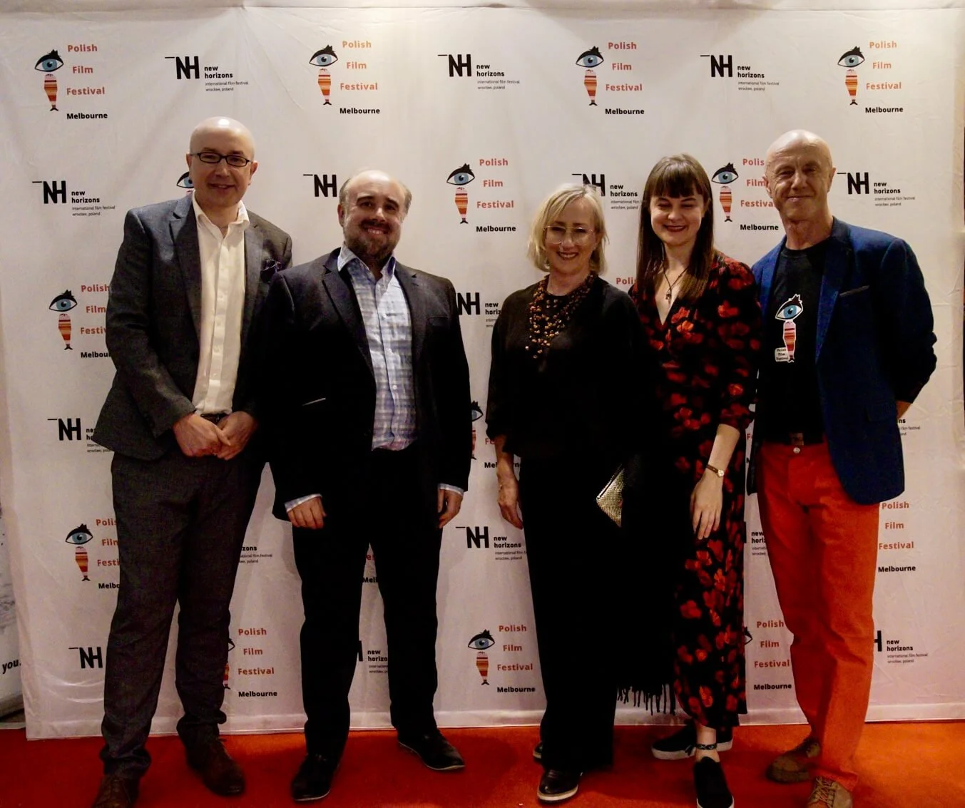 Five people standing in front of a backdrop with the Polish Film Festival Melbourne logo, dressed in formal attire, smiling for a photo at the Polish Film Festival event.