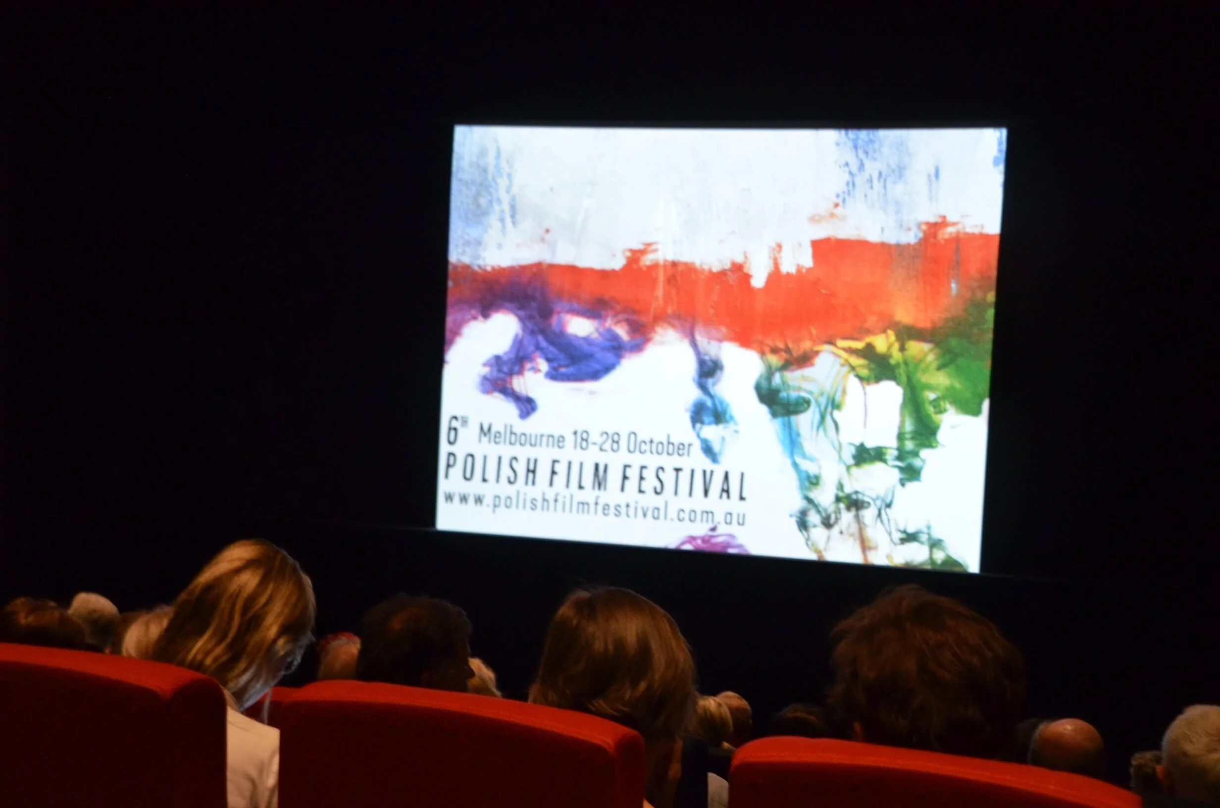 Audience watching a presentation at the Polish Film Festival in Melbourne, displayed on a large screen with colorful abstract artwork and event details.
