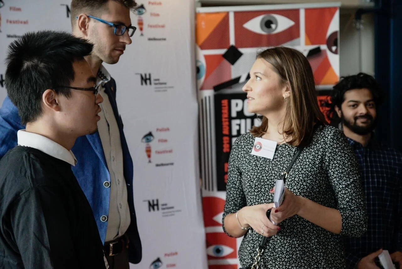 Four people are having a conversation at an event. One woman is talking to two men, with a third man smiling in the background. They are standing in front of a colorful backdrop and a white board with logos.