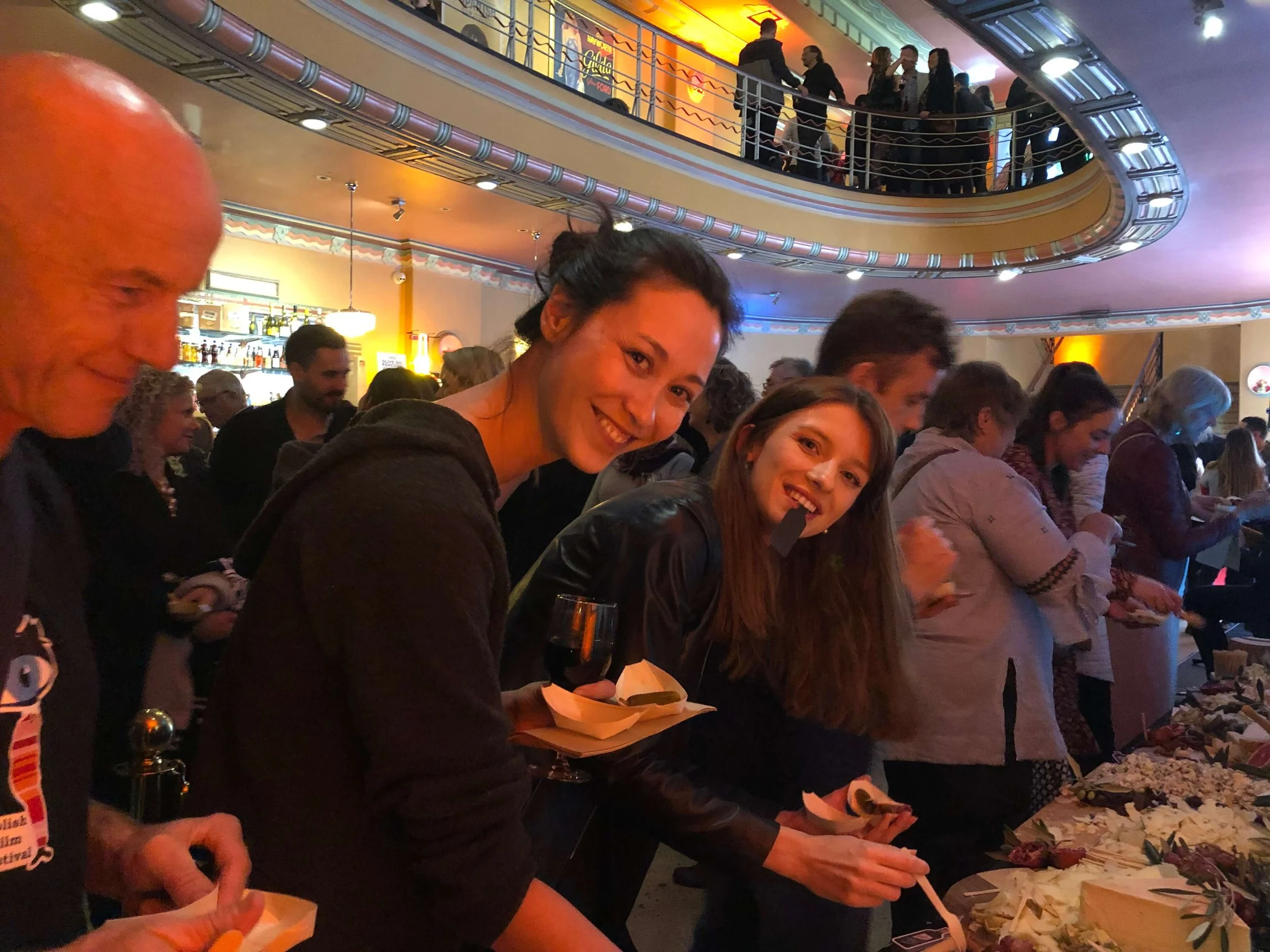 People standing in line at a buffet in a crowded restaurant or event space, with a ceiling balcony and people socializing above.
