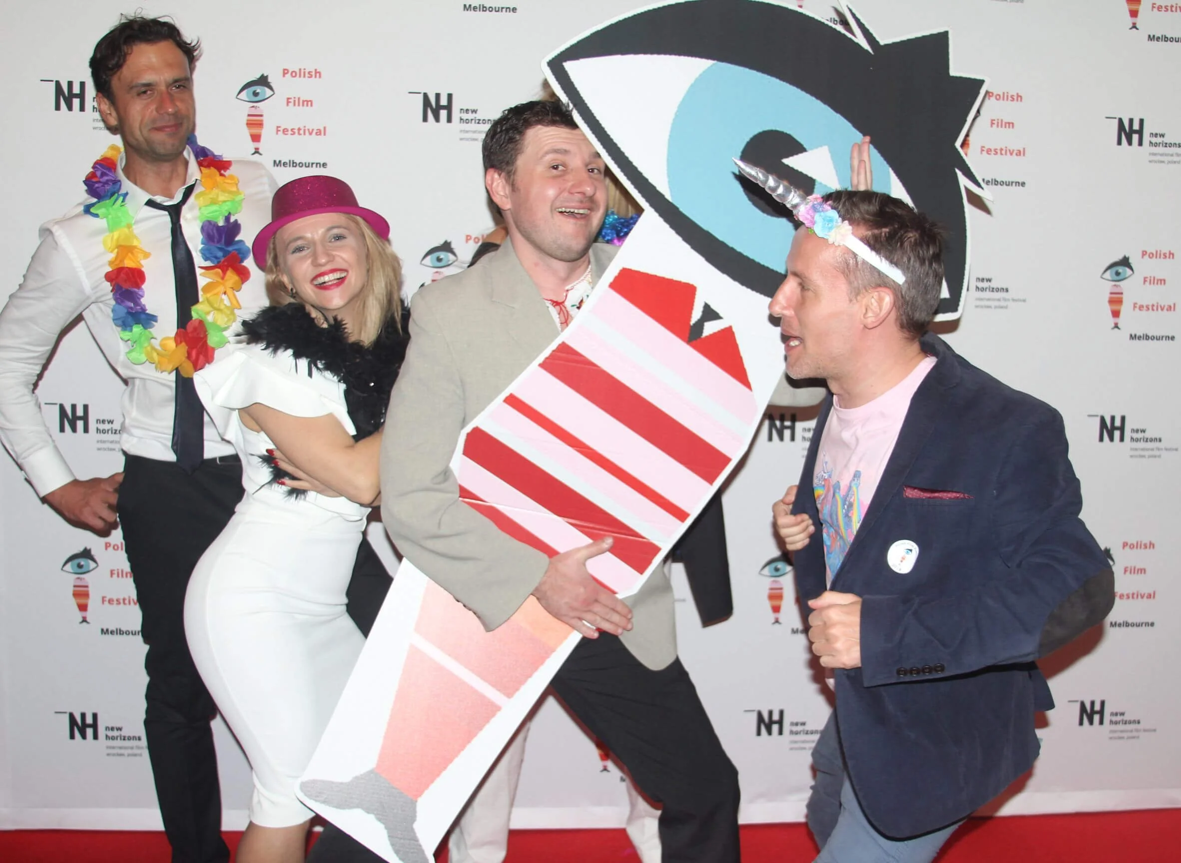 Group of people at a festival posing for a photo, holding a prop shaped like an eye with a blue iris, a rainbow-colored unicorn horn, and red stripes, wearing party accessories like a pink hat and a colorful flower lei, in front of a backdrop with fe
