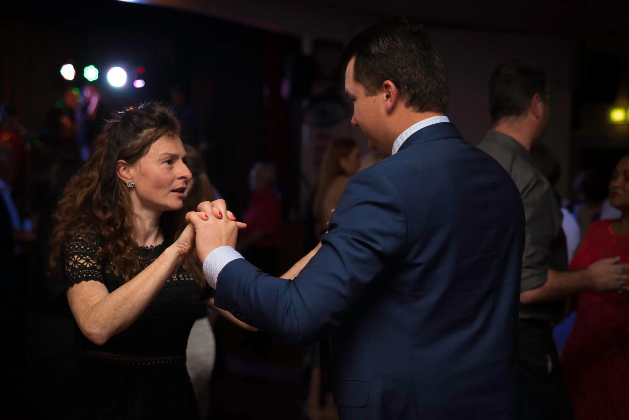 A woman with curly brown hair and earrings and a man in a suit dancing at a social event with other people in the background.
