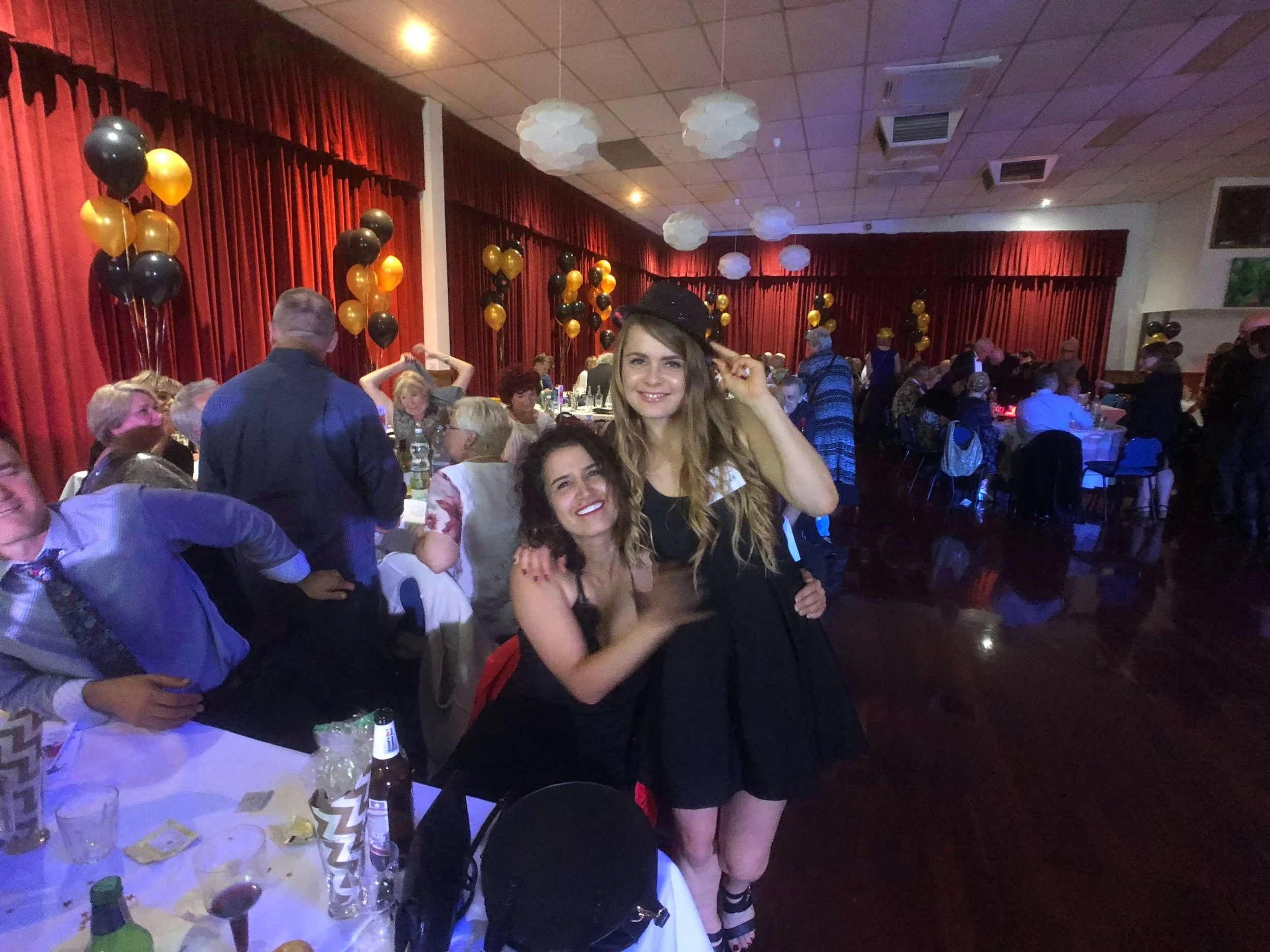A woman smiling and wearing a black hat, standing next to another woman at a party or celebration with red curtains, balloons, and tables in the background.