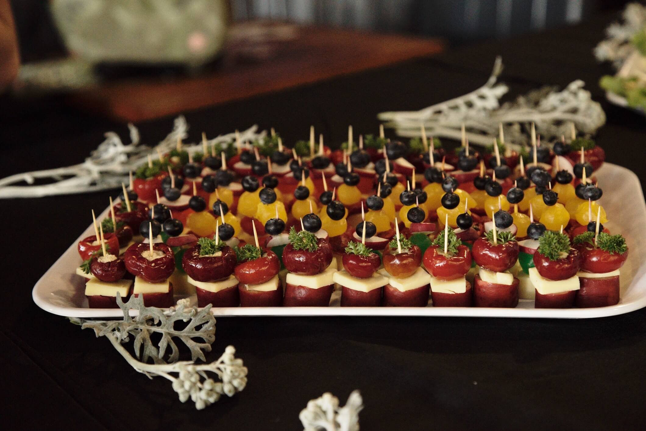 A tray of various colorful skewered appetizers, including cherry tomatoes, cheese, and olives, garnished with greenery, placed on a black table with decorative white branches.