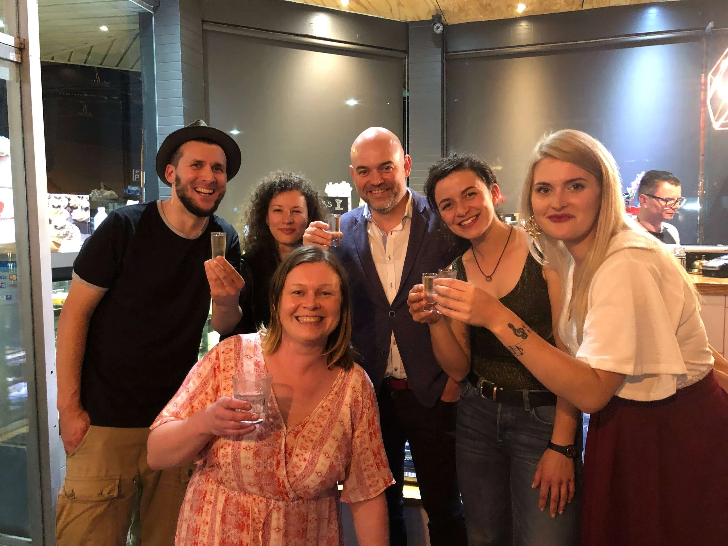 Group of six friends enjoying drinks together in a restaurant or bar, smiling at the camera. A woman in the front holds a glass of water, and others hold small glasses, with some standing and others sitting.