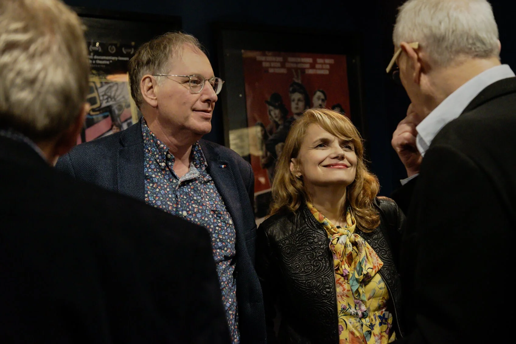 Four people engaged in conversation, two men and one woman smiling, in a room with posters on the wall.