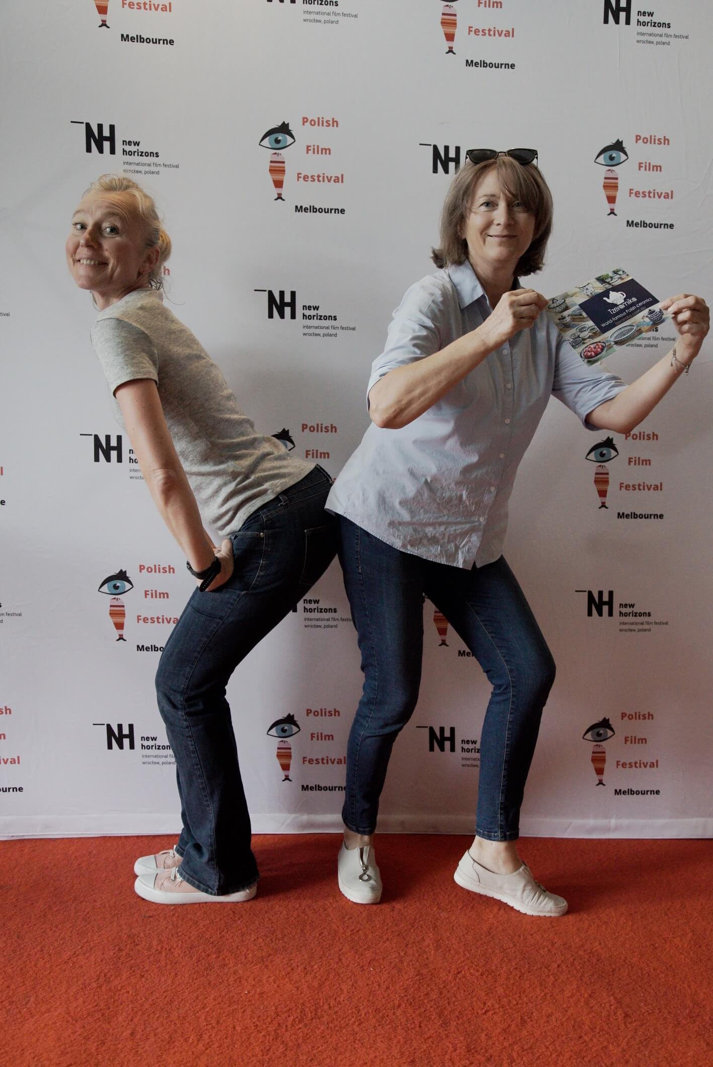 Two women pose back-to-back in front of a banner for the Polish Film Festival in Melbourne. Both are smiling, wearing casual clothes, and holding a brochure. One woman has blonde hair and wears a gray t-shirt and jeans, while the other has brown hair