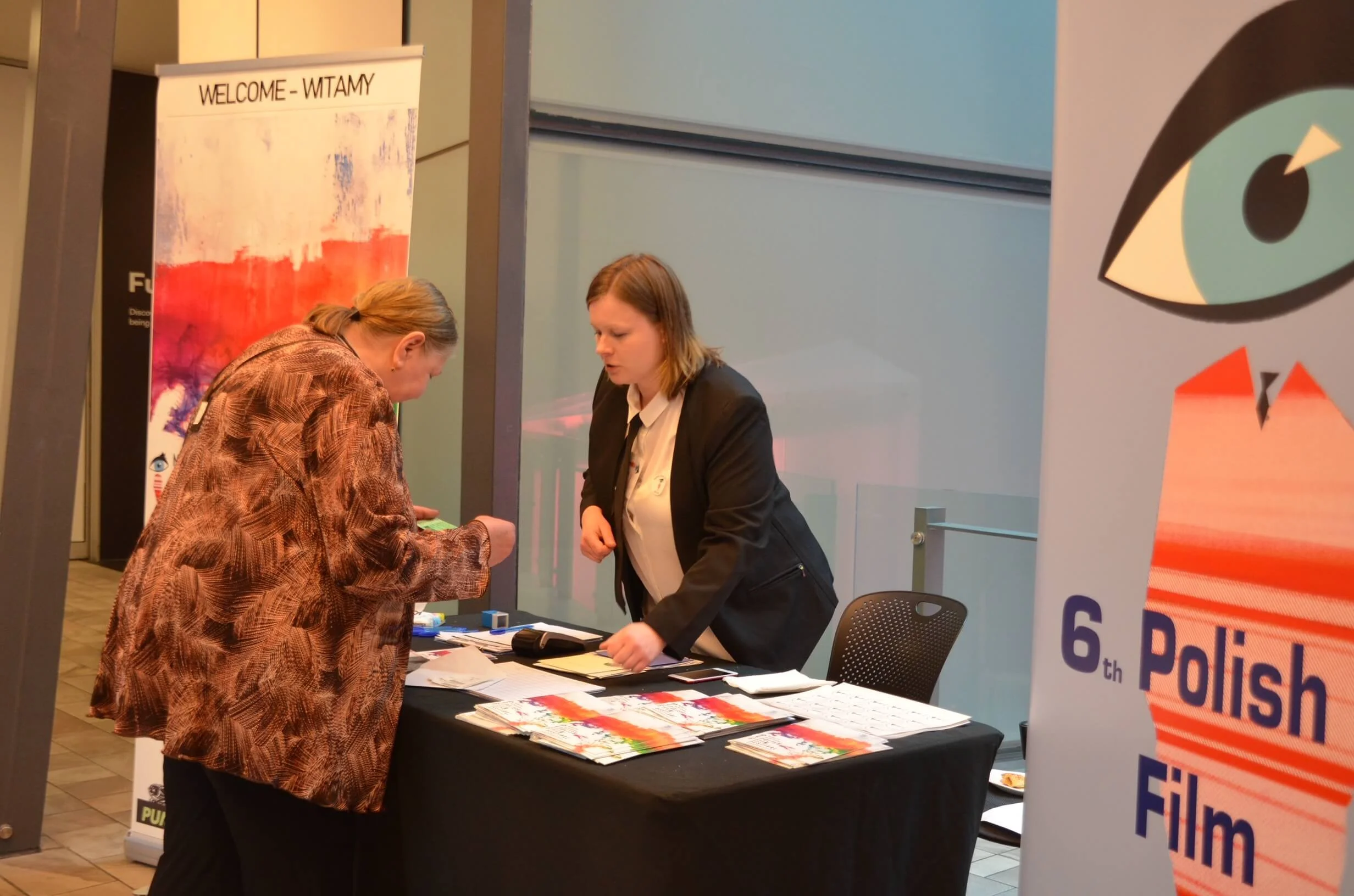 Two women at a registration or information table during an event. One woman is signing or filling out a form, while the other is assisting or answering questions. The table has brochures and papers. There is a colorful banner behind them and a large 