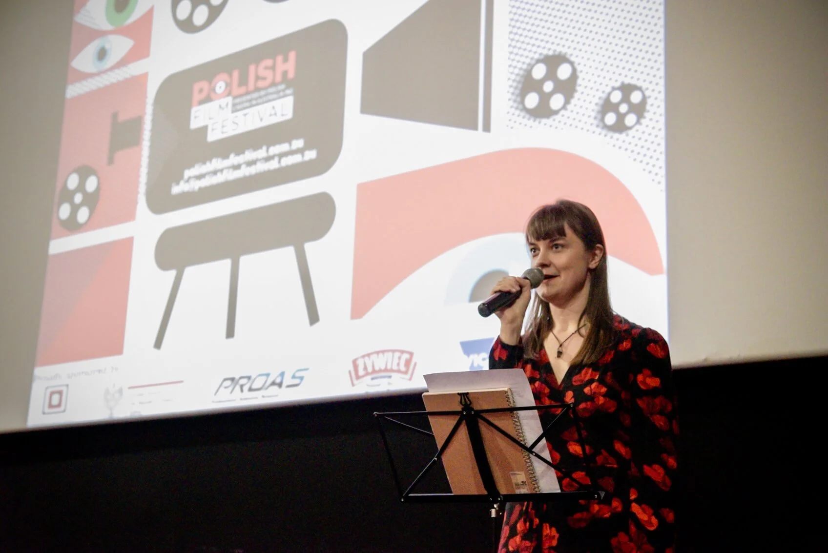 A woman with brown hair and bangs, wearing a black dress with red flower patterns, is speaking into a microphone on stage during a presentation. Behind her, a large screen displays a graphic design with abstract shapes, a chair illustration, and logo