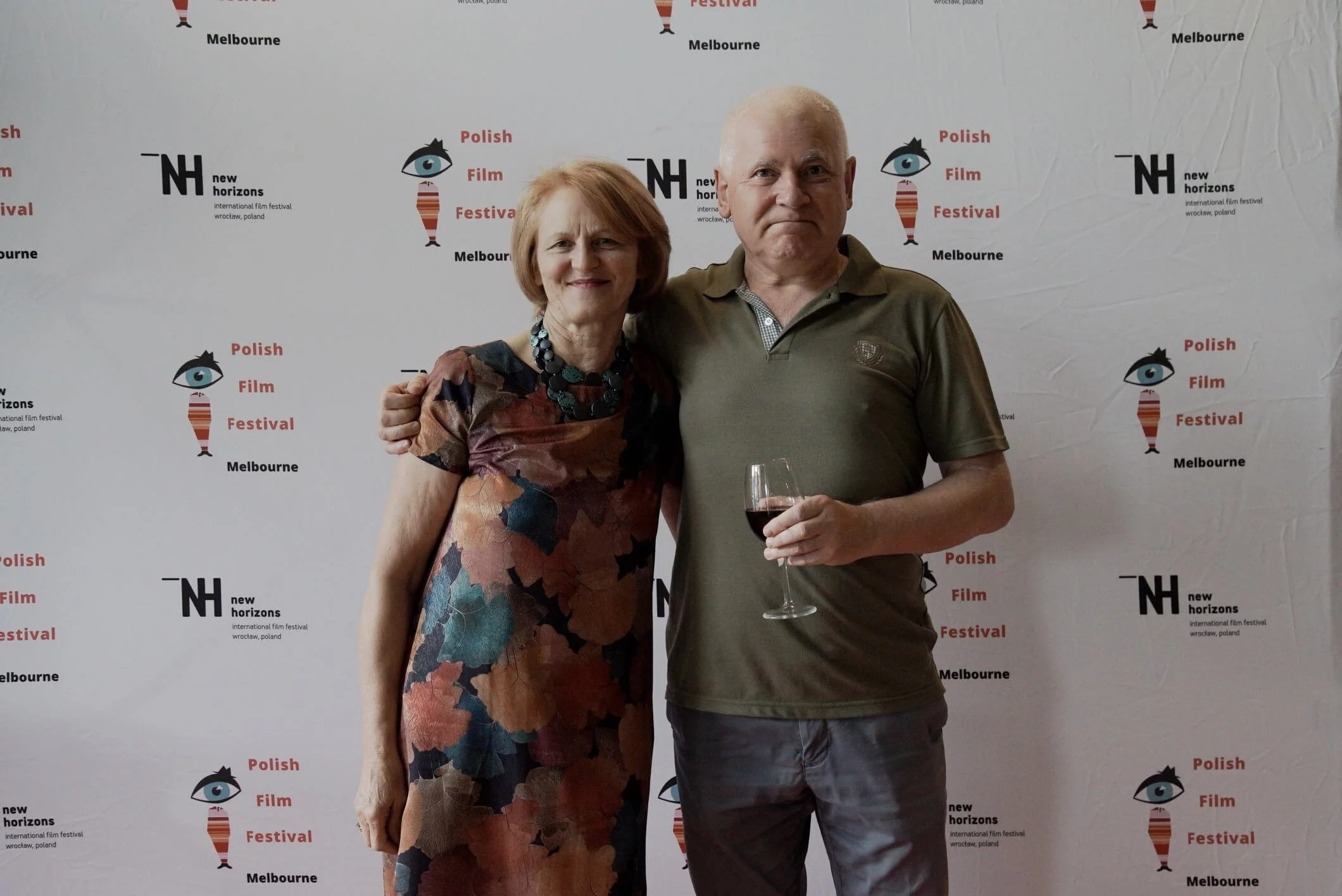 A woman and a man standing together at the Polish Film Festival in Melbourne, Australia. The woman is wearing a multicolored dress and a chunky necklace, and the man is holding a glass of red wine and dressed in a green polo shirt. The background fea