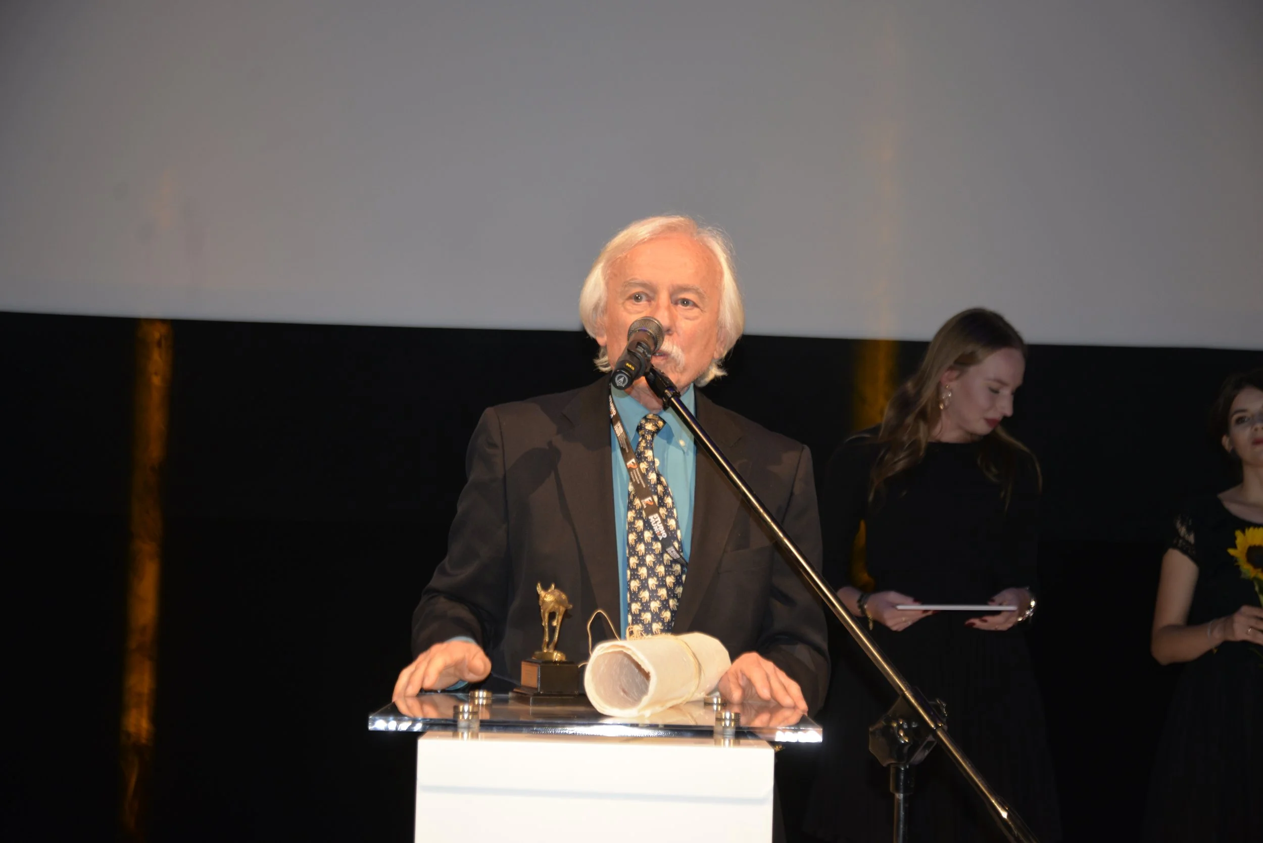 An elderly man with white hair and a mustache speaking into a microphone at an award ceremony. He is wearing a black suit, a blue shirt, and a patterned tie, and is holding a rolled-up document and a small trophy. Two women are standing behind him, o