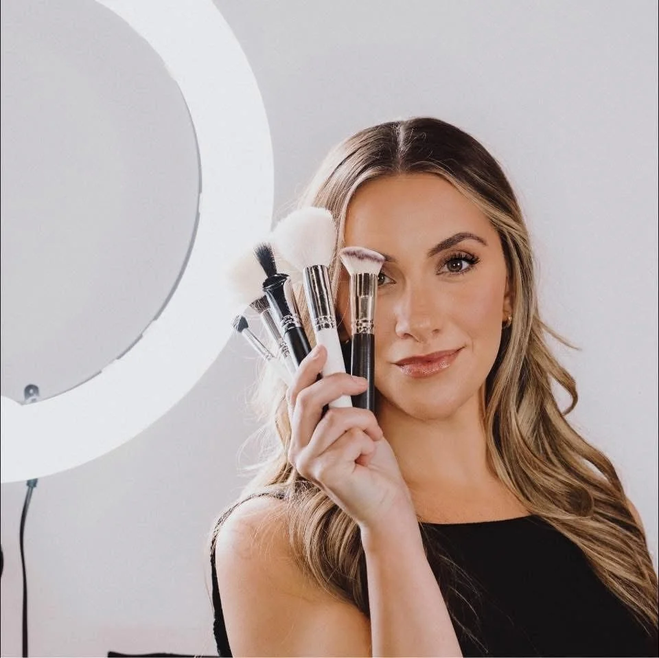 A woman holding makeup brushes near her face, standing in front of a ring light.
