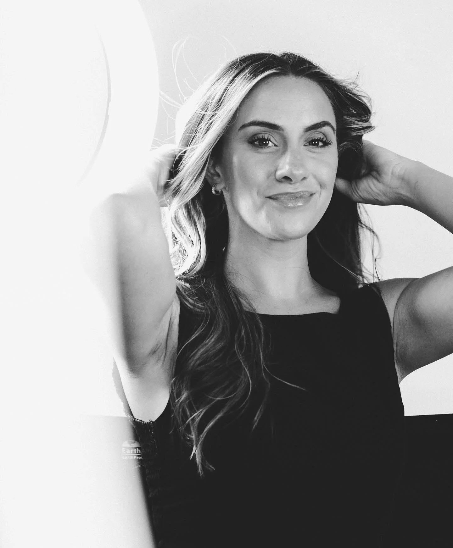 A woman with long wavy hair smiling, holding her hair back with both hands, wearing a sleeveless black top, in black-and-white.