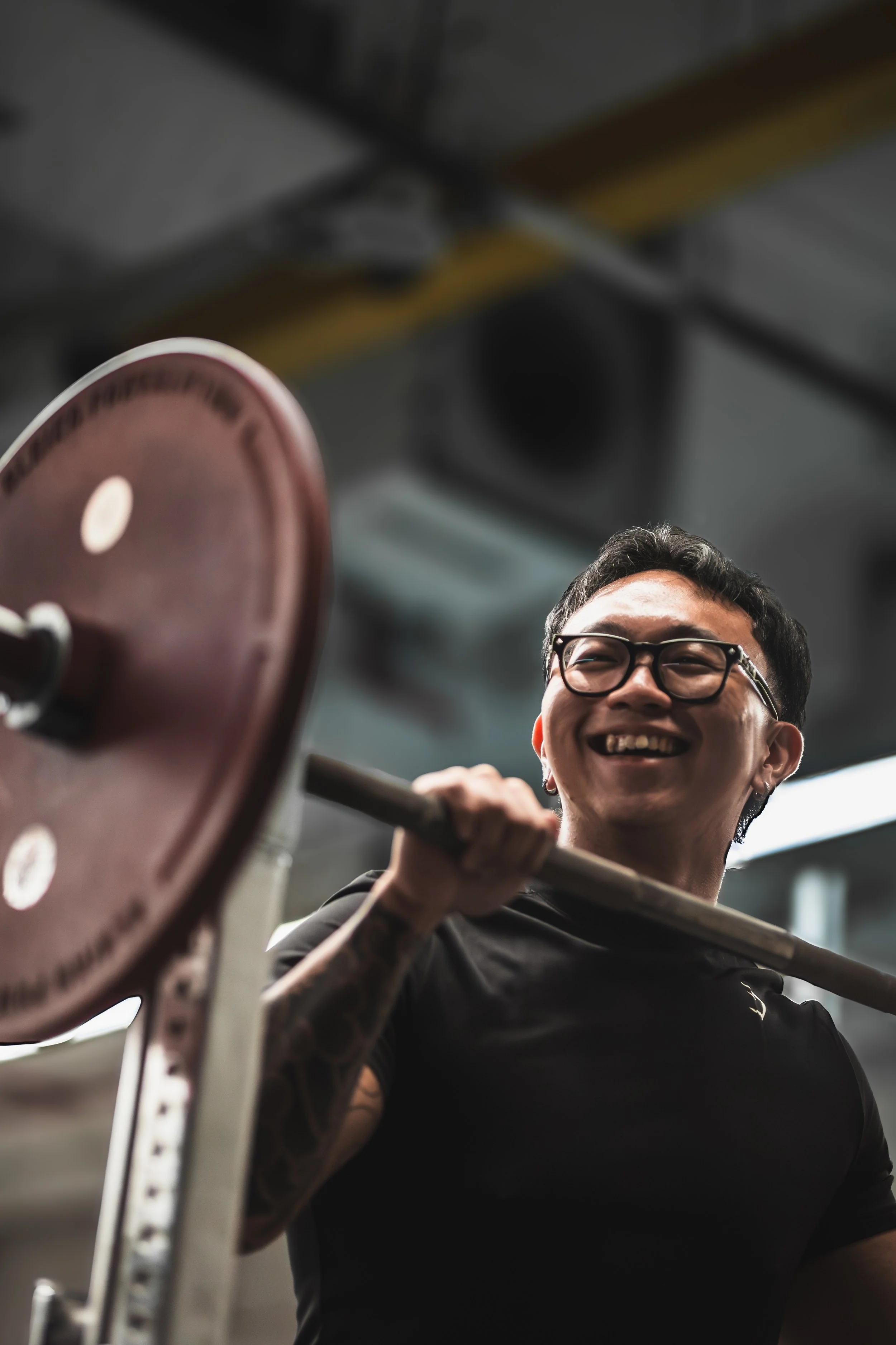 A person with short dark hair, glasses, and a black shirt smiling while lifting a barbell in a gym.