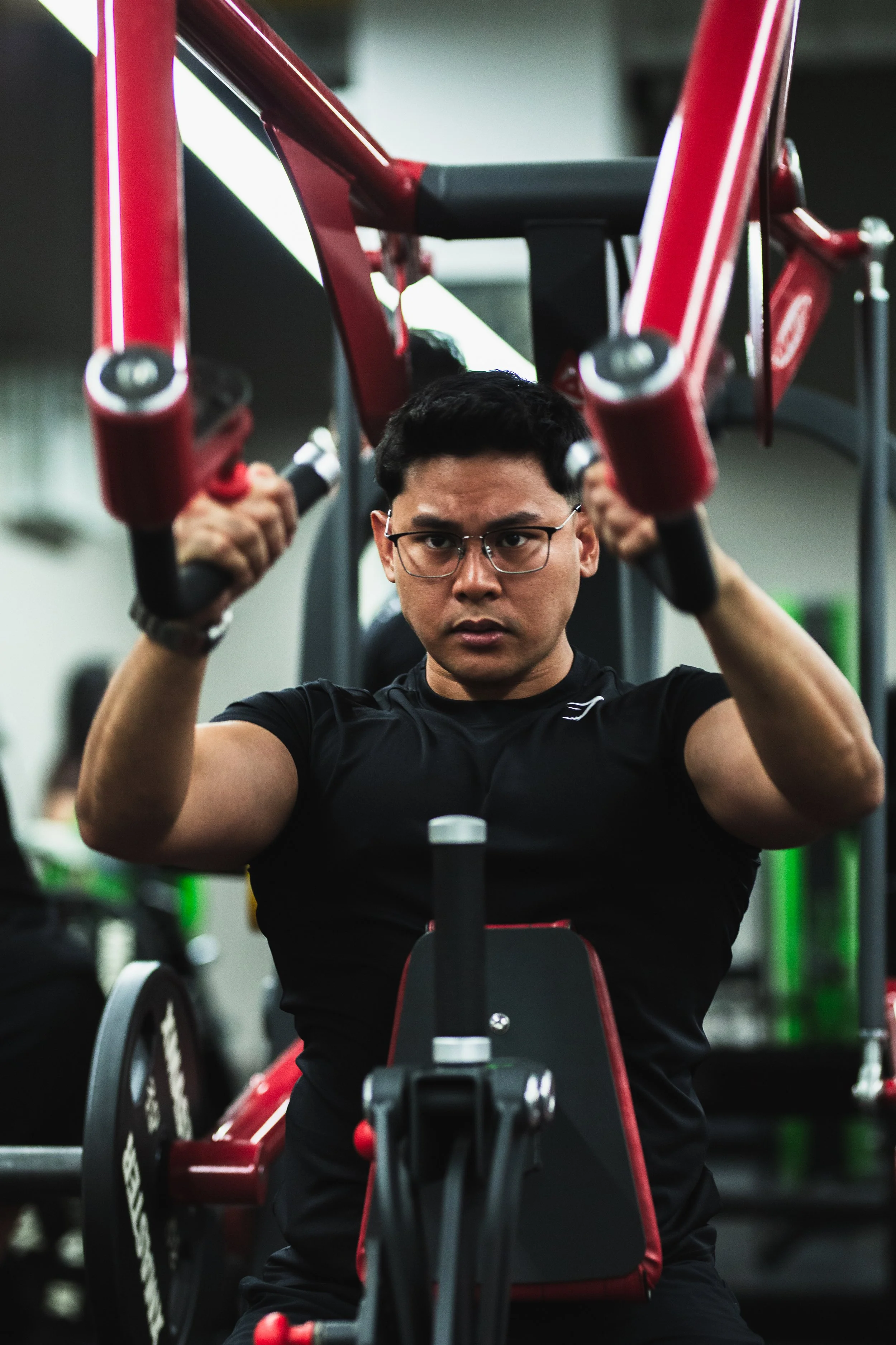 A man in a black athletic shirt and glasses sitting in a gym wheelchair, lifting weights with a focused expression.