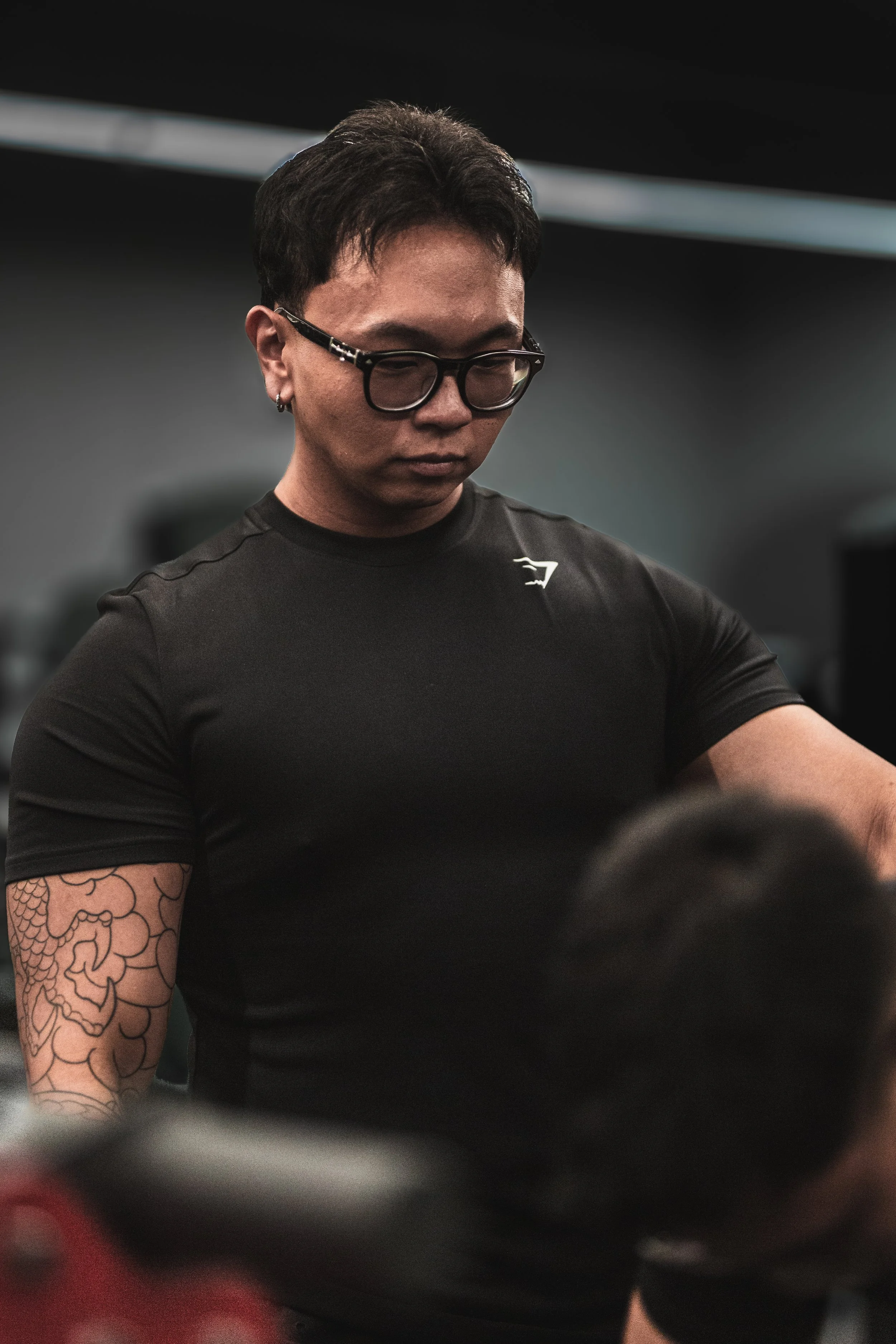 A man with short dark hair, glasses, and earrings, wearing a black shirt with a logo, looking downward, with tattoos on his arm.