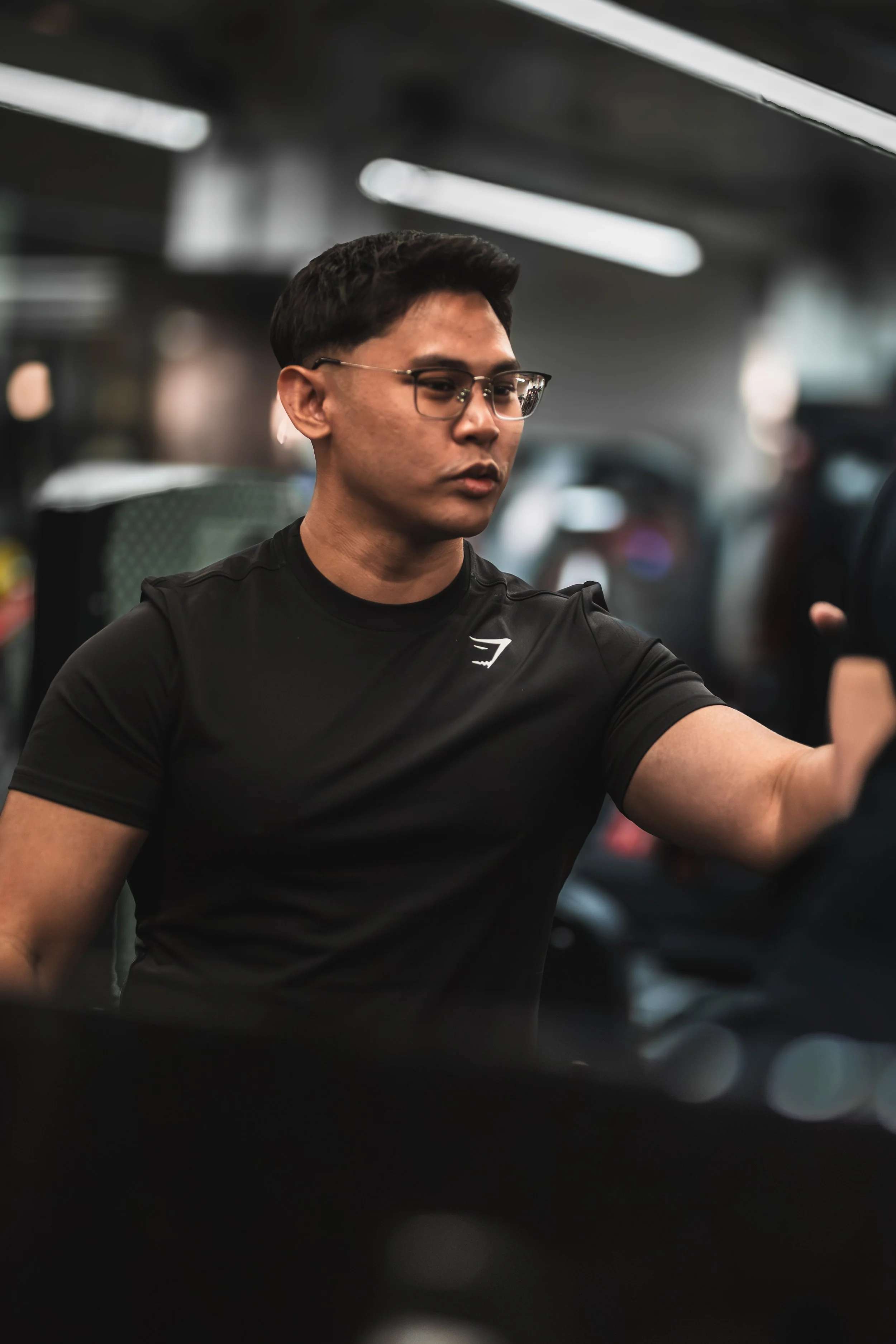 A young man with short dark hair and glasses, wearing a black T-shirt, is in a gym, possibly doing a workout. The background is blurred with gym equipment and lighting.