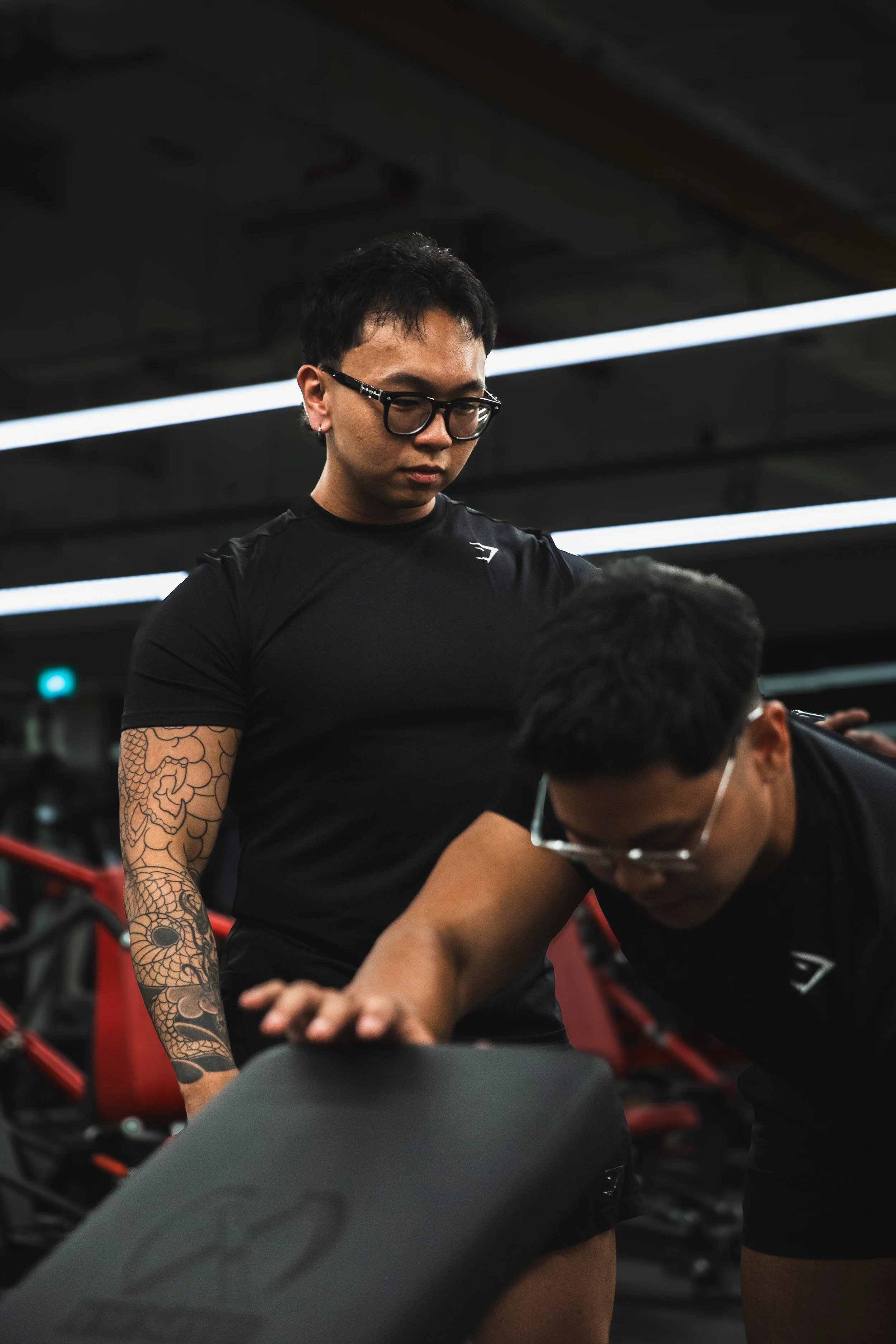 A man with short black hair, glasses, and tattoos on his arm, wearing a black shirt, is assisting another person in a gym or fitness setting with a workout or exercise.