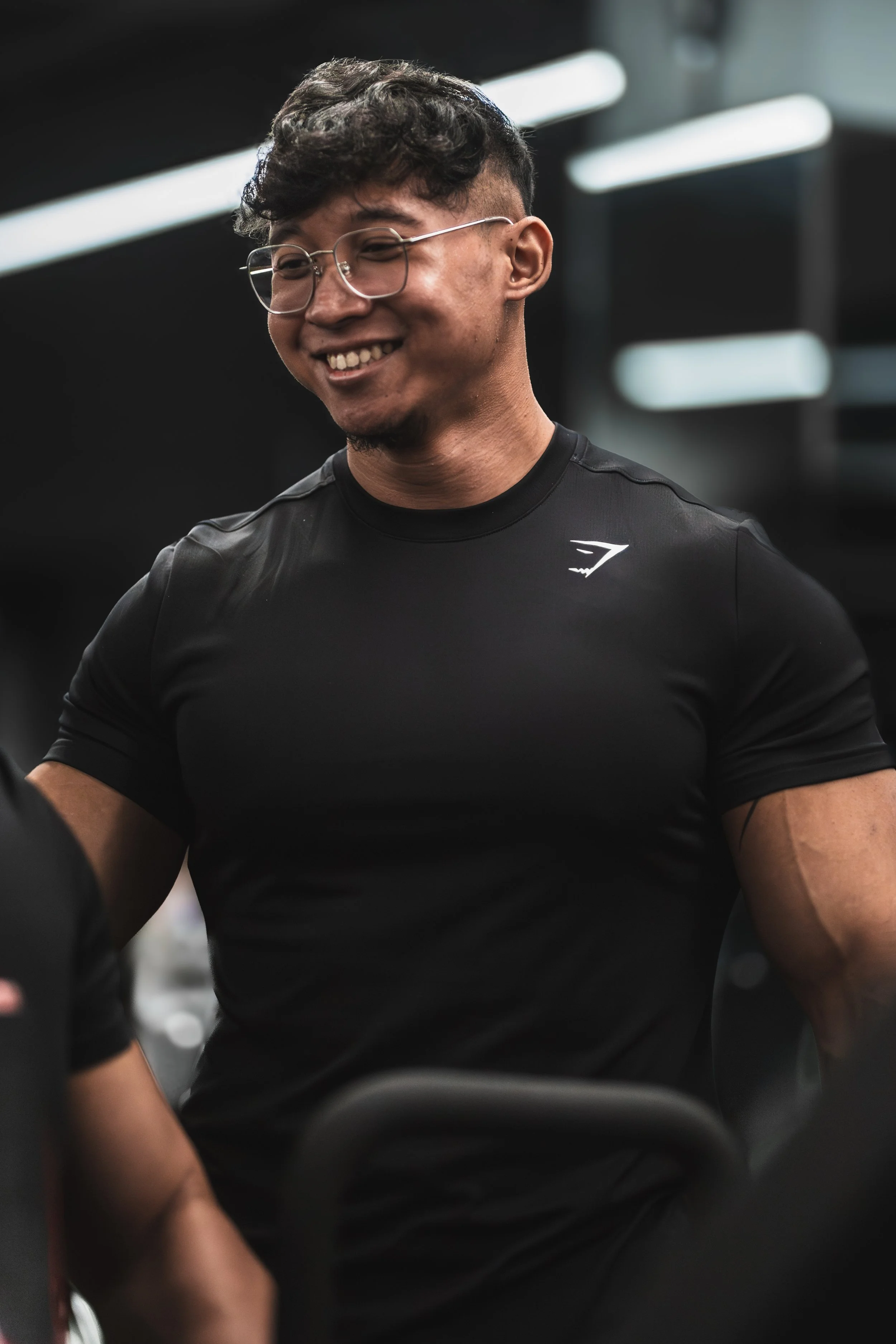 A smiling man with glasses and curly dark hair, wearing a black T-shirt with a small logo, standing indoors with blurry background lights.