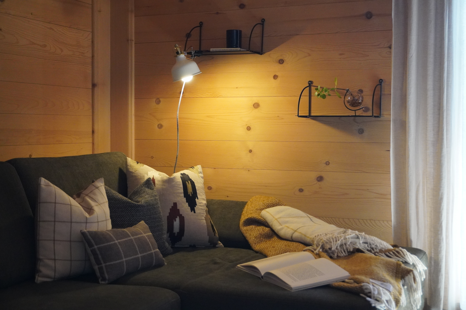 Gemütliches Wohnzimmer mit grauer Couch, mehreren Kissen, einer offenen Buchseite, einem gelben Kissen, einem Plaid, einer stilvollen Holzverkleidung und Wandregalen mit Pflanzen und Dekorationsgegenständen, neben einem Vorhang, der das natürliche Licht im Raum einfängt.