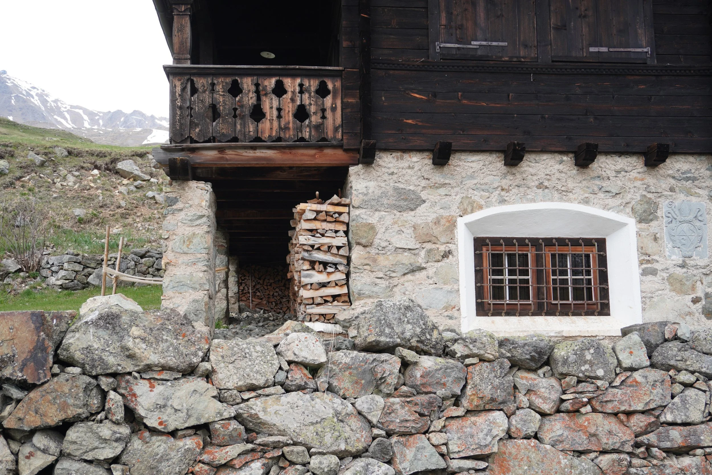 Ein altes Haus mit einer steinernen Foundation, dunklem Holzbau und einem kleinen Balkon aus Holz. Im Vordergrund sind zahlreiche große Steine und ein Holzstapel. Im Hintergrund sind schneebedeckte Berge sichtbar.