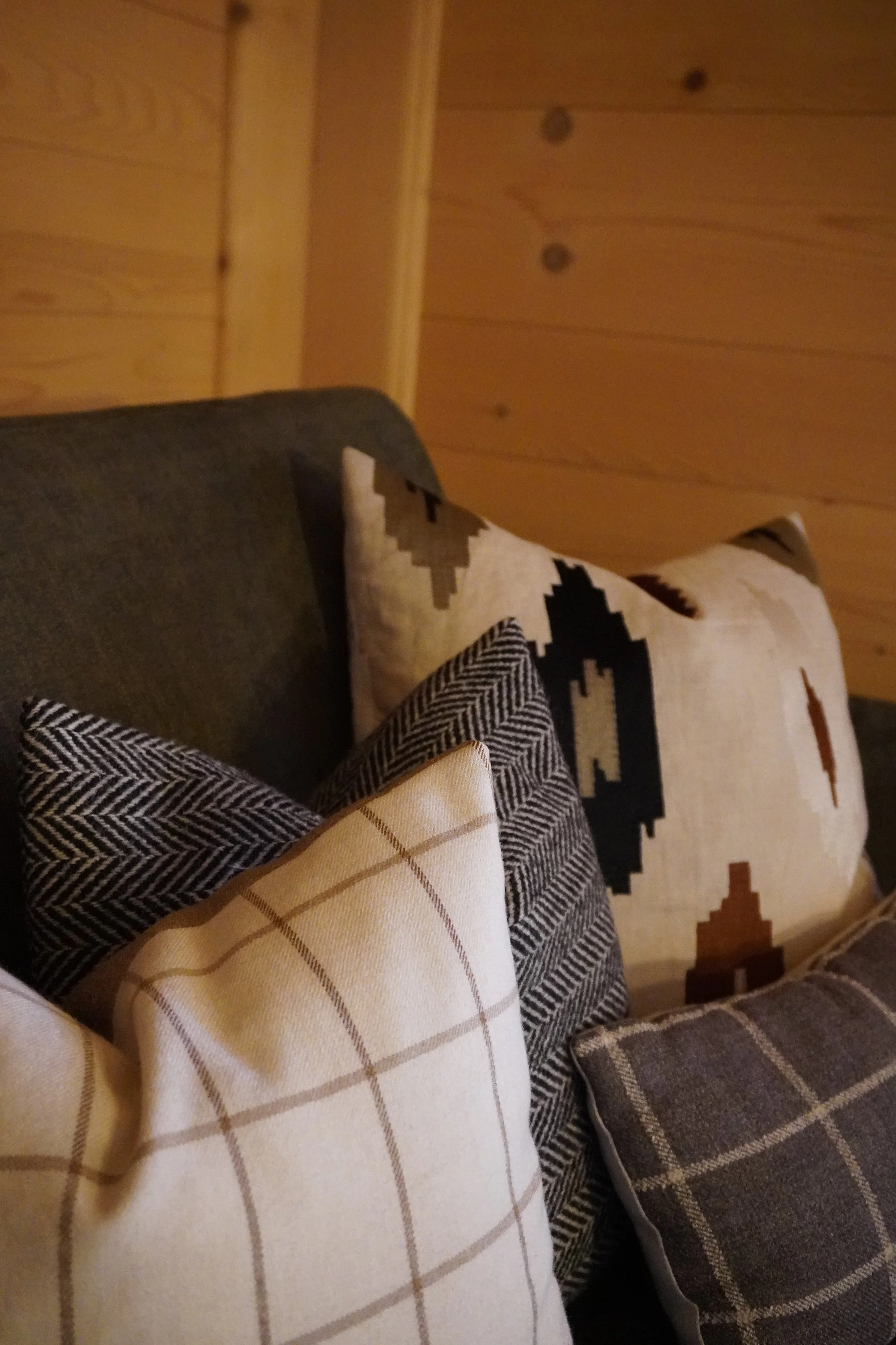 Verschiedene Kissen und Decken auf einer Couch vor einer Holz-Wand.