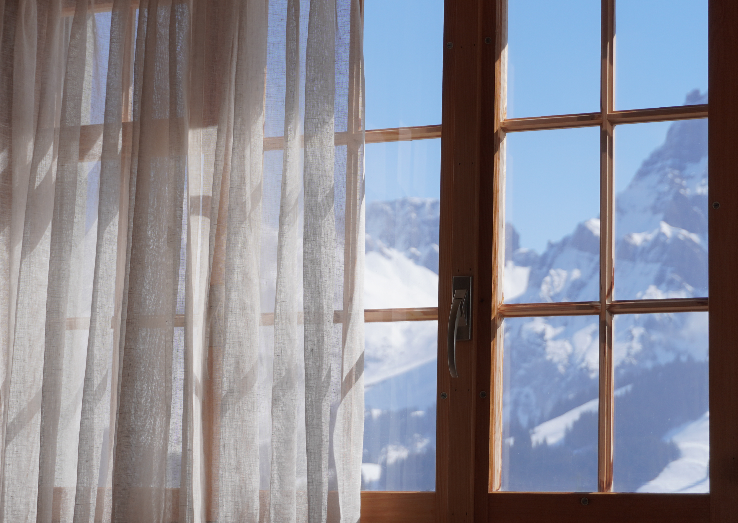 Blick durch ein Fenster mit Holzrahmen auf schneebedeckte Berge und blauen Himmel, Vorhang im Vordergrund.