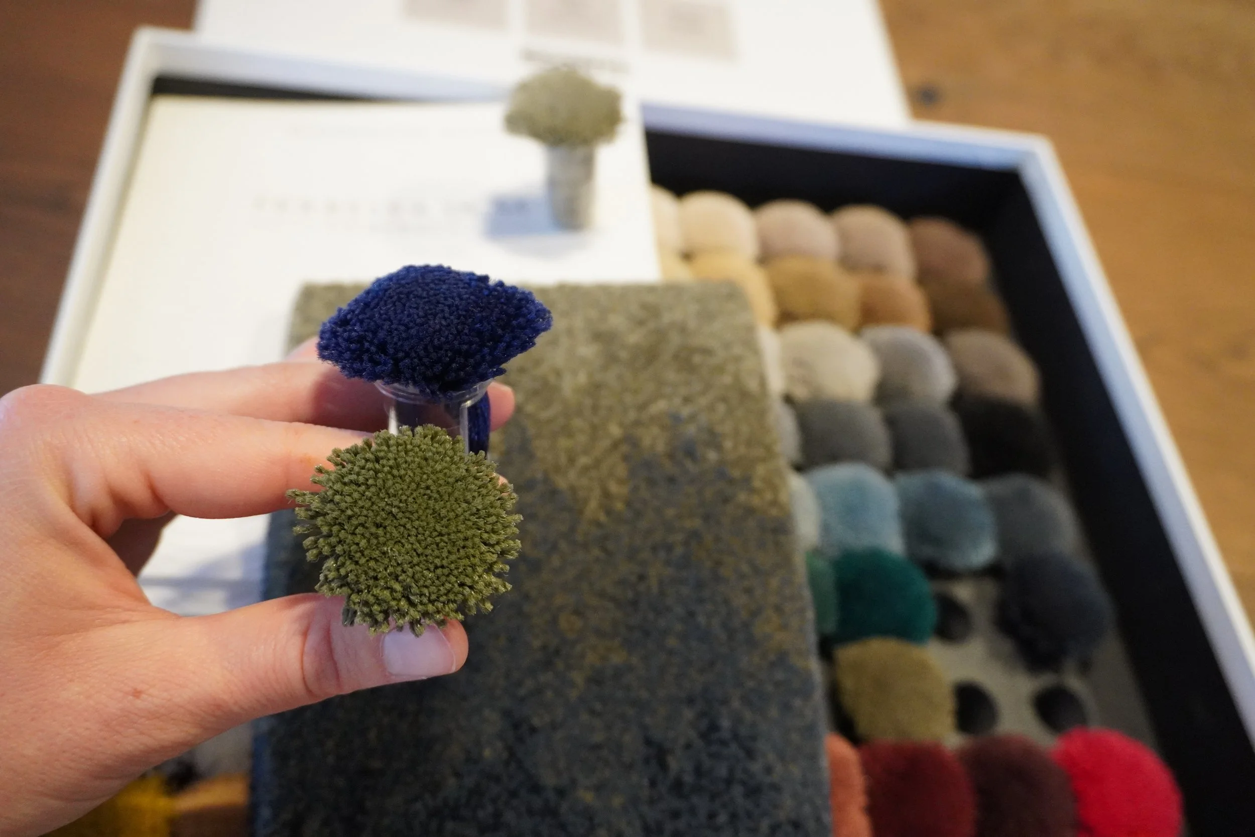 Hand hält drei Wollknäuel in den Farben Blau, Grün und Grau. Im Hintergrund sind weitere Wollknäuel in verschiedenen Farben in einer Kiste sichtbar.