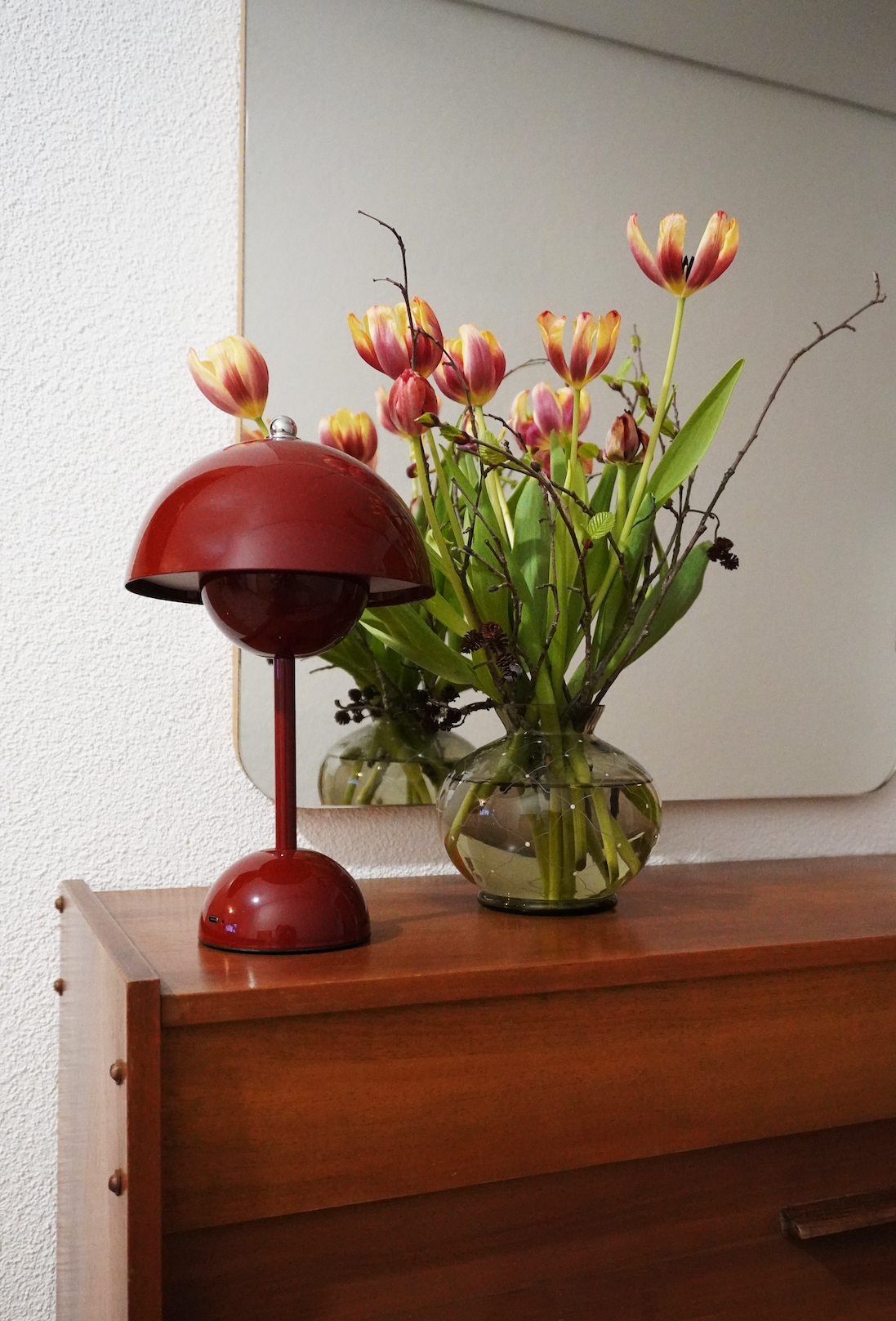 Eine braune Holzkommode mit einer Glasschale voller Tulpen und getrockneter Zweige. Daneben steht eine rote Tischlampe.