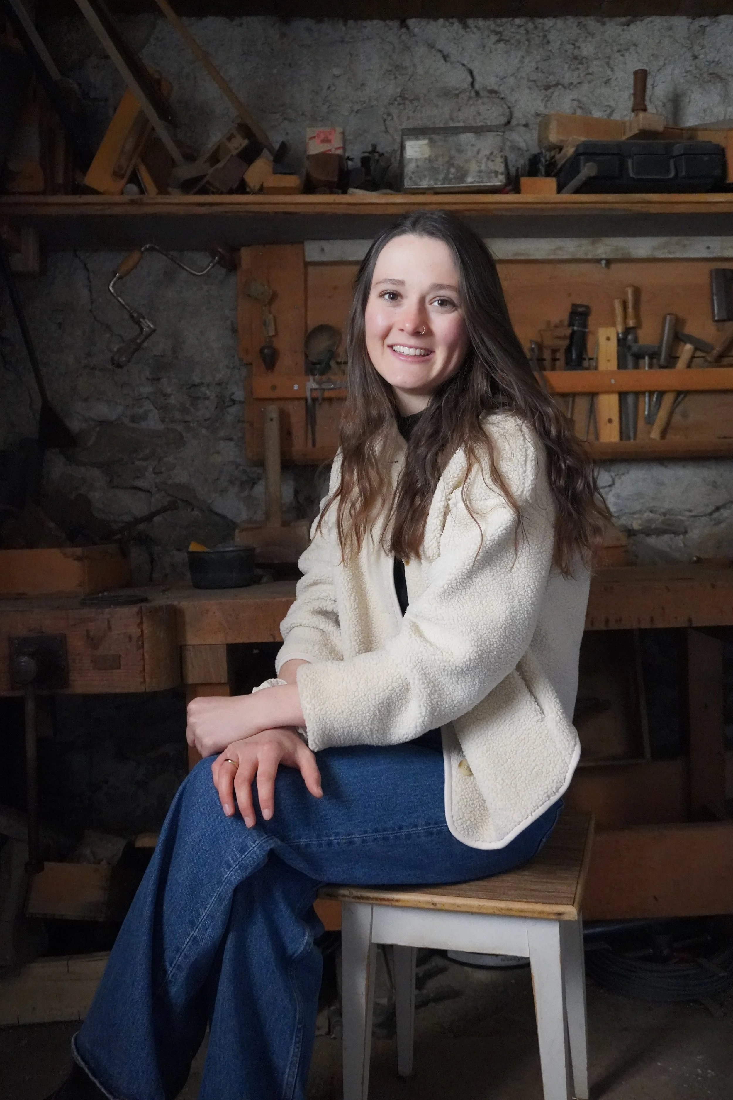 Corinne Gempeler, Inhaberin, sitzend auf einem Stuhl in einer Werkstatt mit Werkzeugen an der Wand, trägt eine weiße Jacke und blaue Jeans.