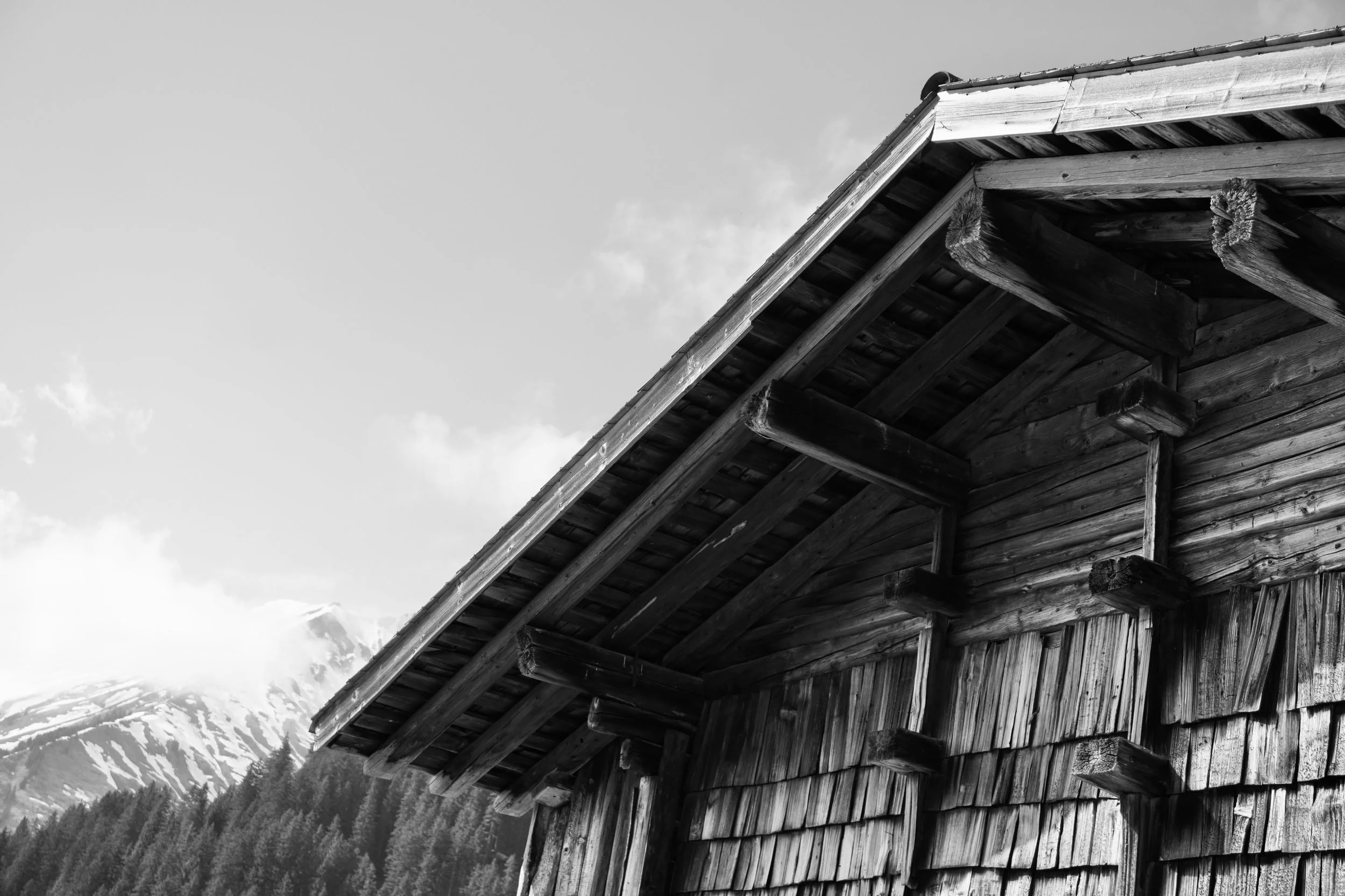 Schwarz-weiß-Foto eines Holzhauses mit steilem Dach, umgeben von Bergen und Wäldern, Wolken im Himmel.