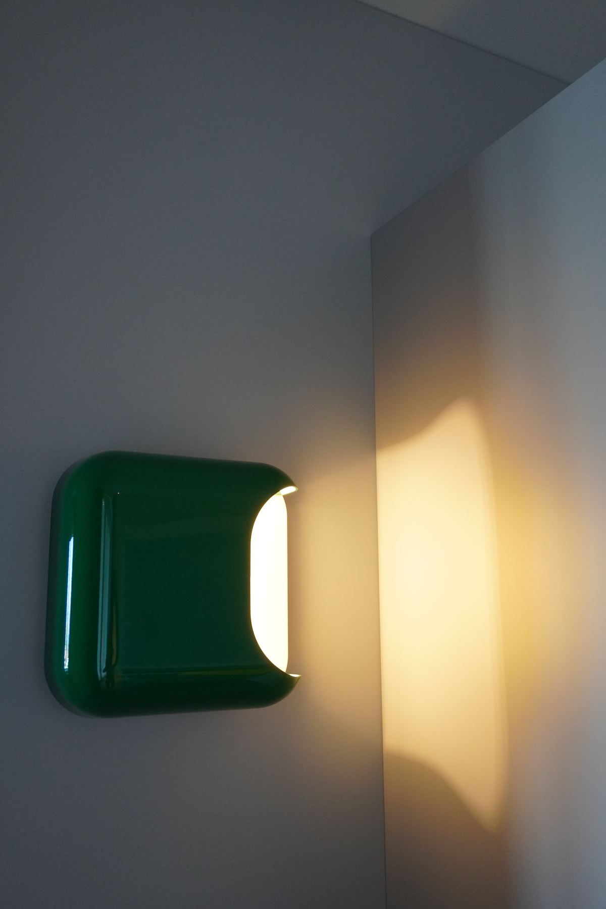 Ein modernes, wandmontiertes Licht mit grünem Gehäuse, das warmes Licht auf eine weiße Wand wirft.