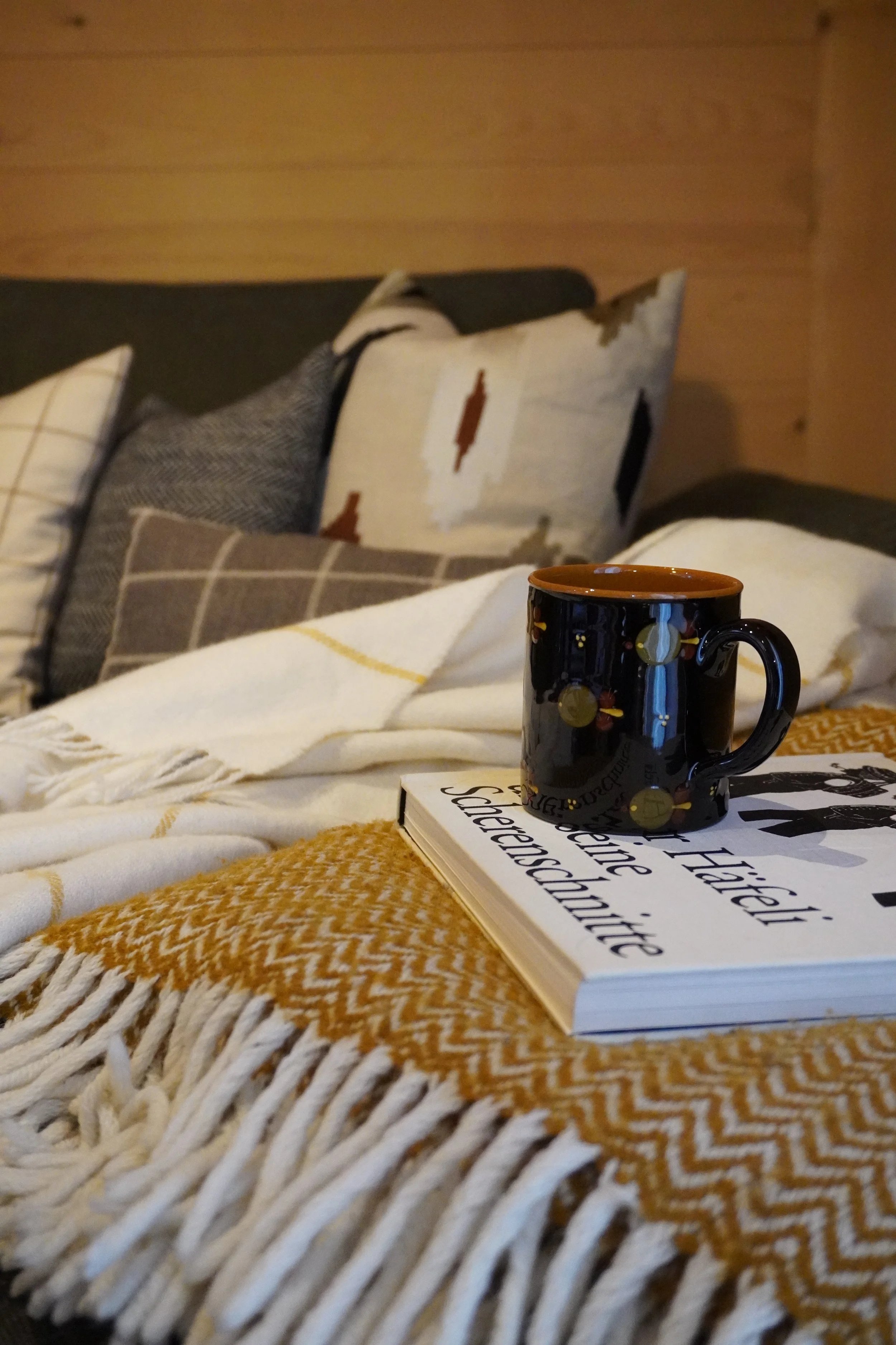 Ein schwarzer Kaffeebecher mit bunten Punkten, der auf einem gestreiften Buch liegt, auf einem gelb-orangenen Deckenstück.