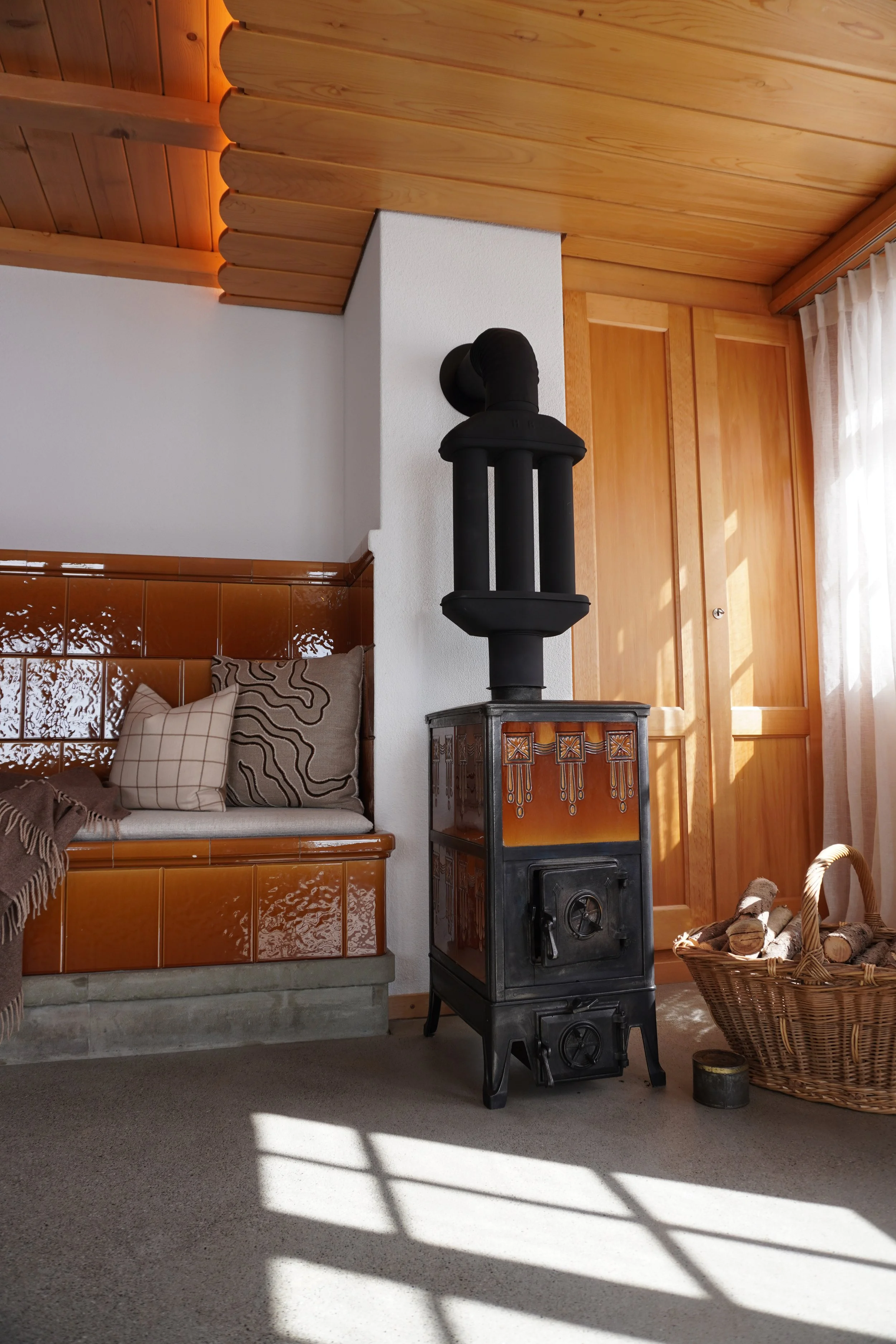 Ein rustikales Wohnzimmer mit Holzdecke, Holzvertäfelung und einem Keramikofen, einem Korb mit Holzscheiten und einer Couch mit Kissen