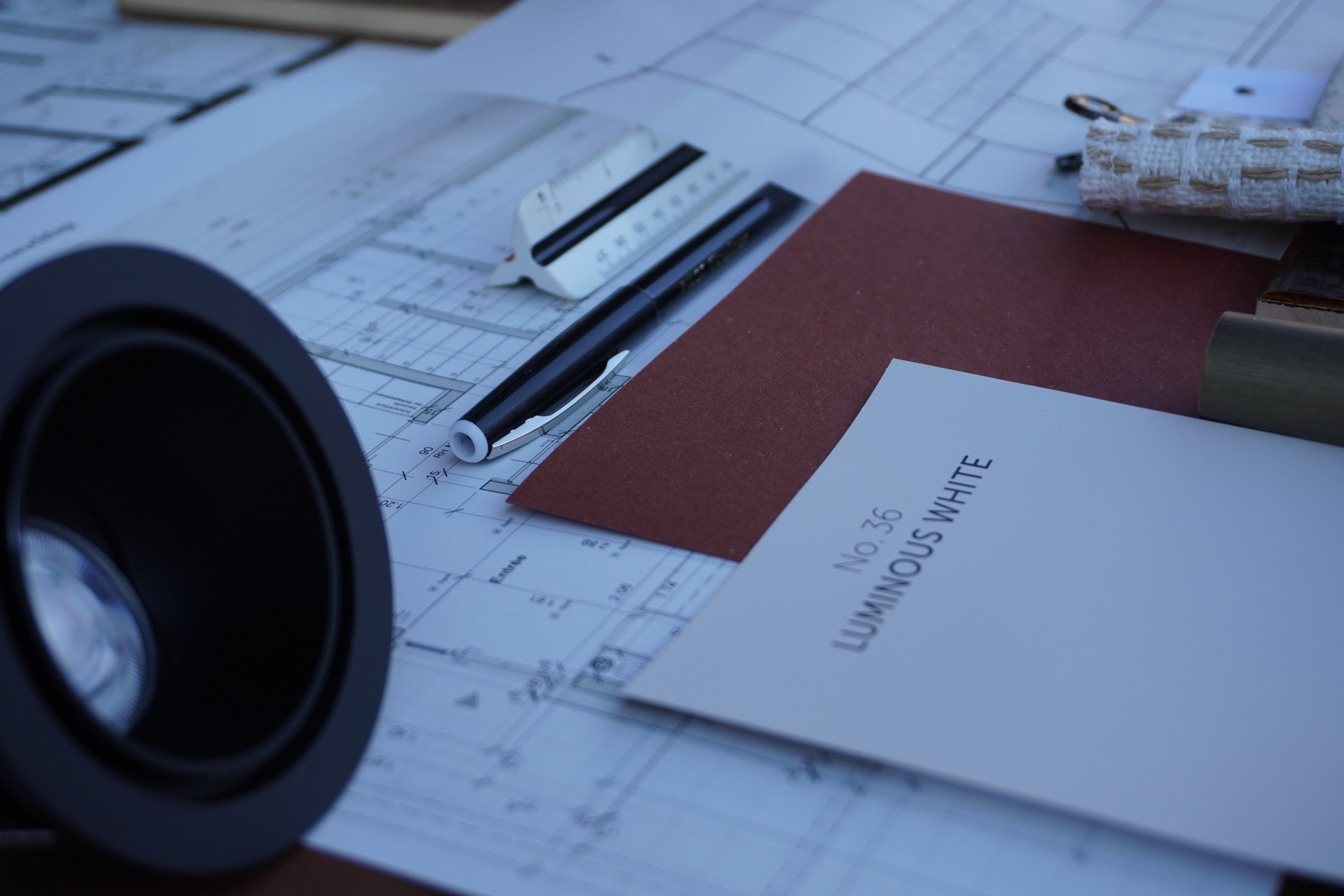 Architekturpläne, ein Cuttermesser, ein Stift, Farbmuster und andere Werkzeuge auf einem Tisch, vermutlich bei der Planung eines Bauprojekts.